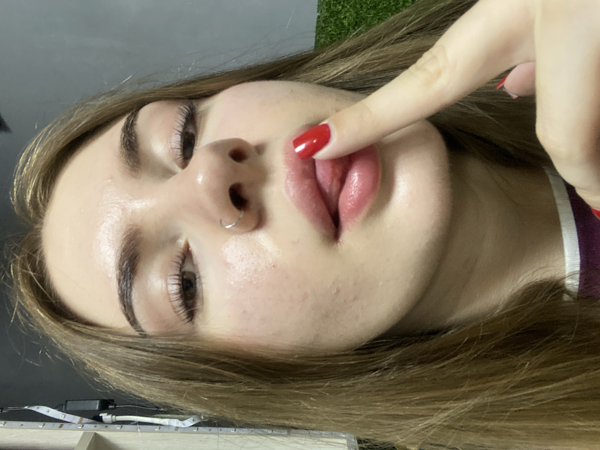 A close-up portrait shows a young woman positioned at an angle, her head tilted to the side. Her face is centered, with her long, light brown hair flowing around her. She has fair skin, a silver hoop nose ring, and dark, long eyelashes on her eyes, which are partially closed or looking downward. Her lips are parted, revealing her tongue, and her right index finger, adorned with bright red nail polish, is touching her upper lip. The skin on her face shows some natural texture, including visible pores and slight blemishes.
In the background, a rough, green textured surface, resembling artificial grass, is visible in the upper right. The lower left foreground reveals a white LED light strip with an attached black control box or power brick. A portion of a garment with white and purple/maroon stripes is visible on her right shoulder/neck area. The lighting highlights her skin, giving it a somewhat dewy appearance. There is no visible text in the image. The overall scene suggests a casual, perhaps self-portrait, with a focus on facial features and a playful pose.