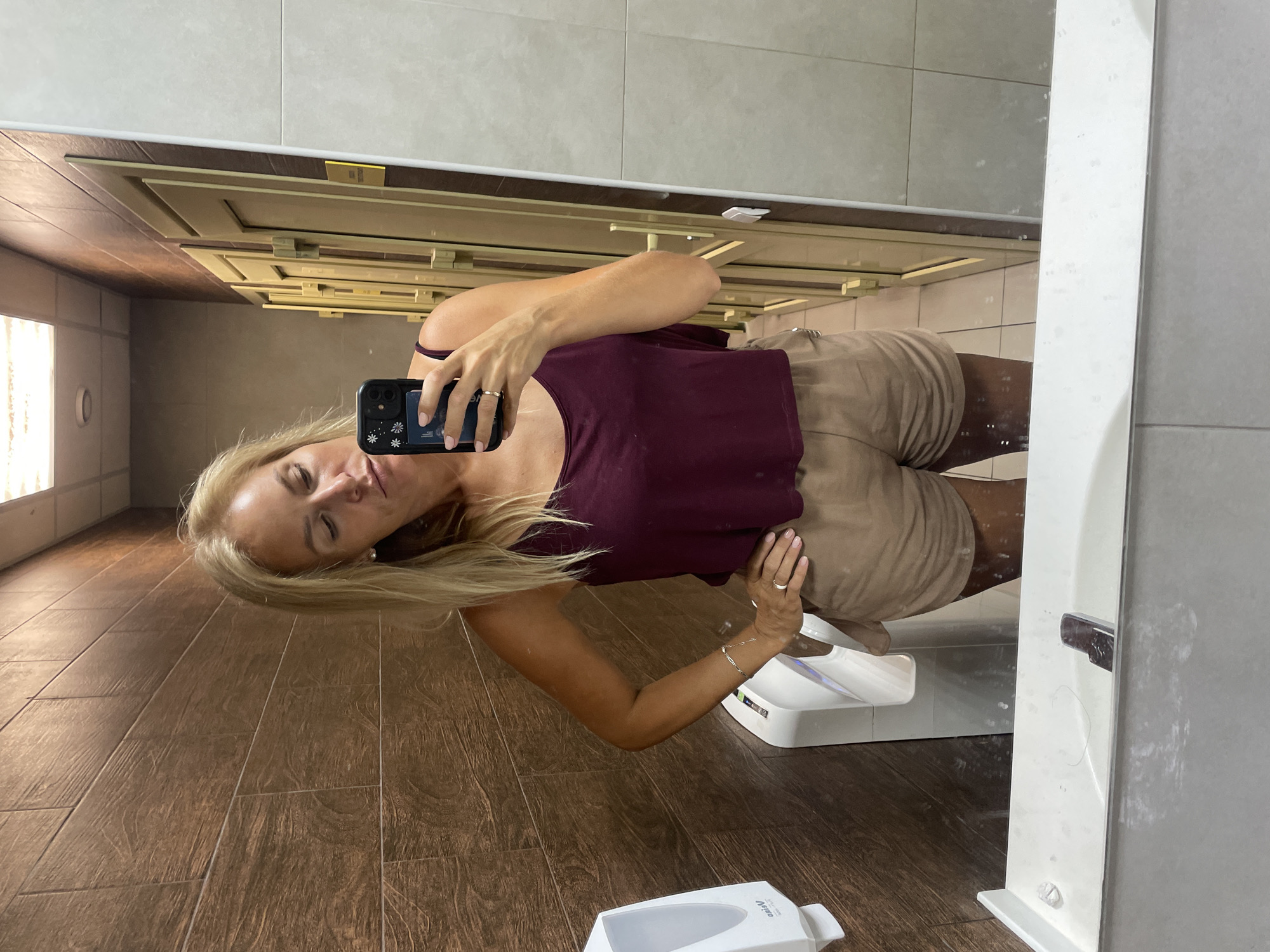 A woman with blonde hair takes a selfie in a restroom. She is wearing a maroon tank top and beige shorts. She has a ring on her left ring finger and a bracelet on her left wrist. Her left hand is on her hip. In the background, there is a brown tiled wall, a white hand dryer, and several doors. The woman is holding a black cell phone with a floral case in her right hand, facing the mirror. The sink is visible in the foreground, and the floor is tiled. The lighting appears to be artificial. The location is identified as a public restroom in the city of Agoy, Russia.