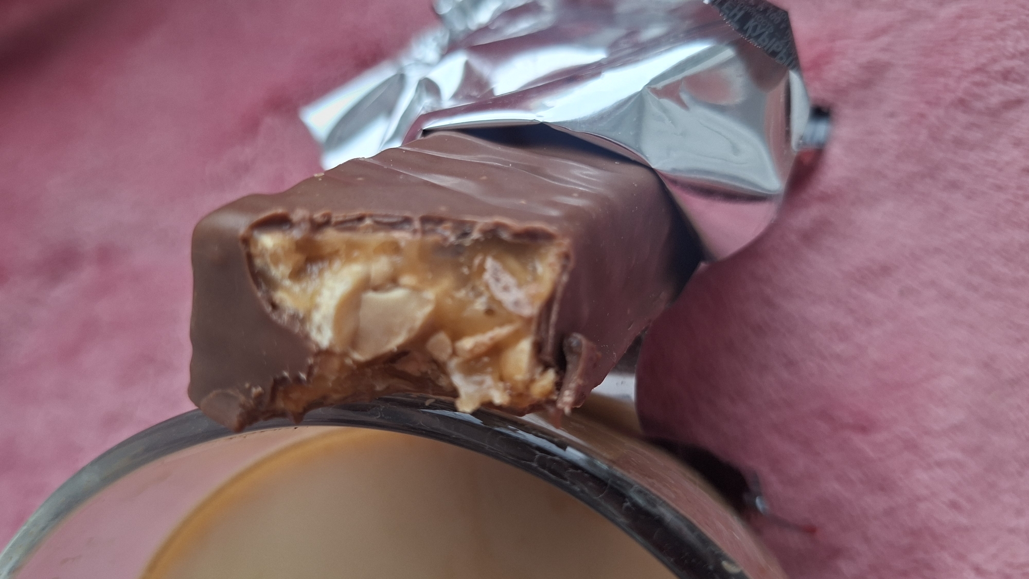A partially unwrapped chocolate and nut candy bar with a bite taken out of it rests on the rim of a glass. The candy bar wrapper is silver. The background features a blurred, reddish-pink textured surface. The location is Забалоцкі сельскі Савет, Belarus.