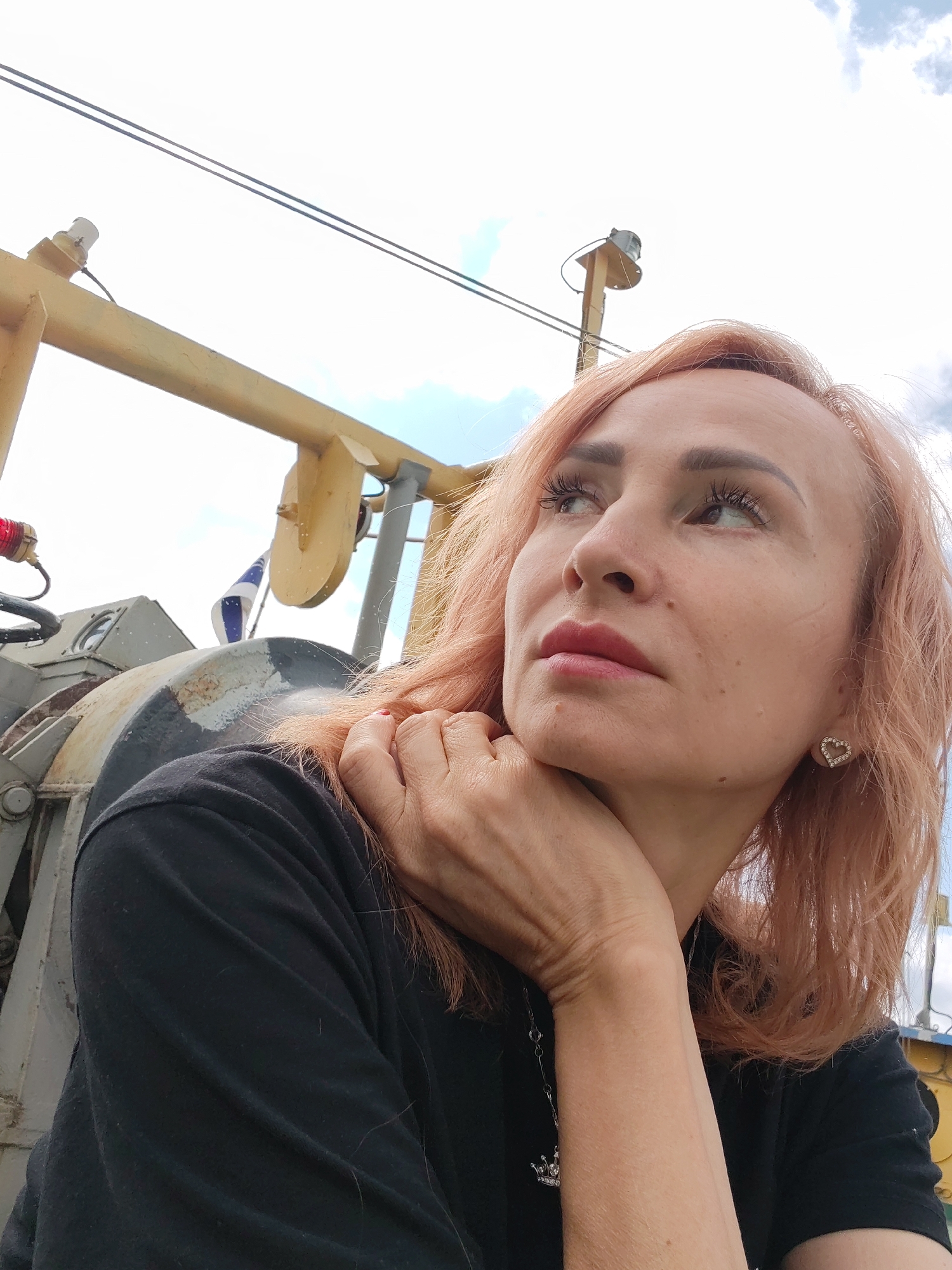 A woman with light pink hair is captured in a close-up, low-angle shot outdoors. She is wearing a black shirt and a silver necklace with a crown pendant. Her left hand is resting on her chin and neck, and she is looking upwards and to the right. A small, heart-shaped earring with rhinestones is visible on her right ear. In the background, there is industrial-looking equipment, including a yellow structure and a grey cylindrical object with a flag attached. The sky above is partly cloudy. The image is described as being taken in Kostyaevka, Russia.