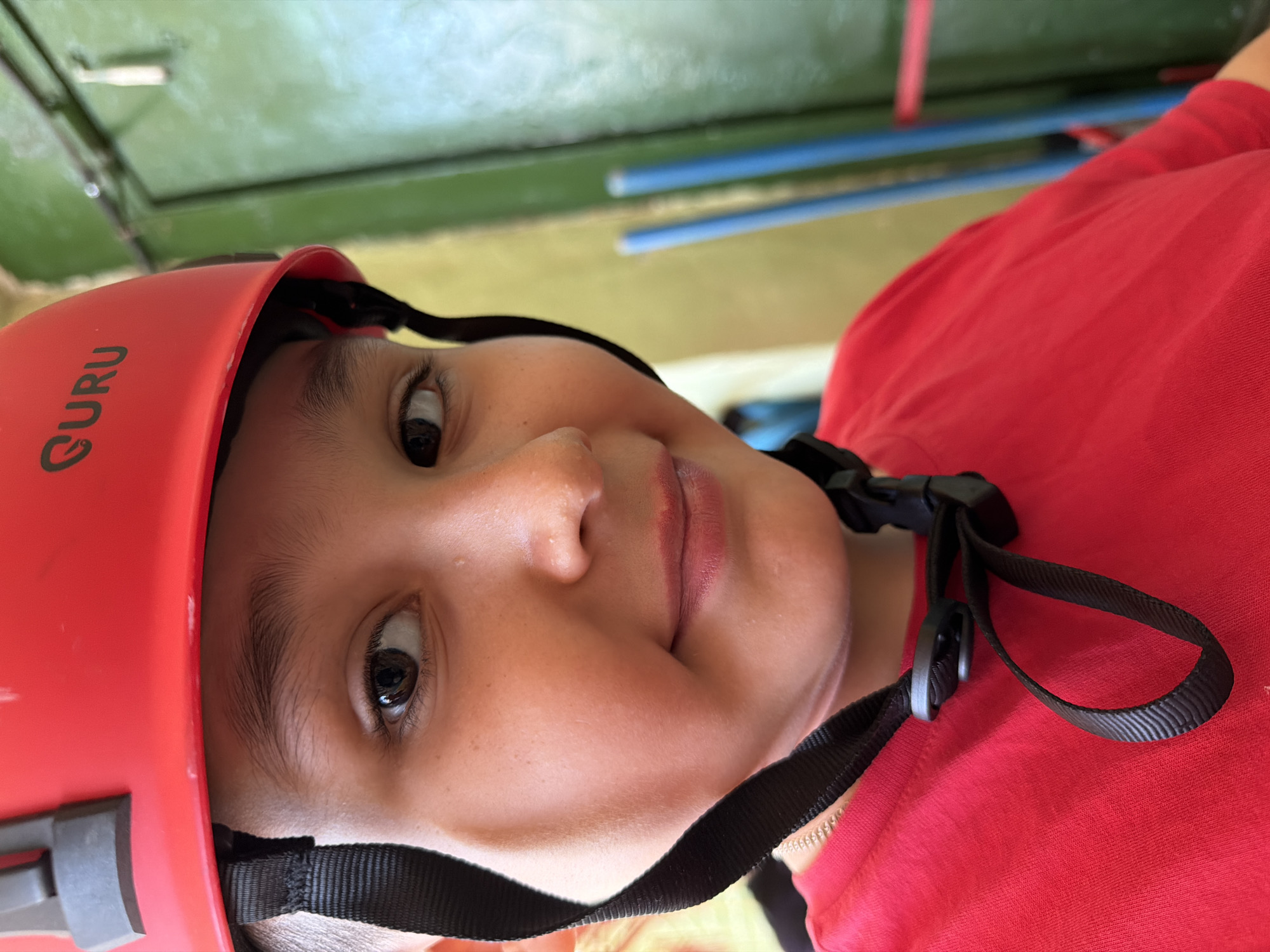 A person wearing a red helmet with the word "Guru" on the left side is the primary subject of the image. The helmet has black straps that are fastened under the person's chin. The person is wearing a red shirt. The background consists of green and yellow surfaces with blue cylindrical objects in the distance. Without more information, I cannot identify the location in Izhevsk, Russia.