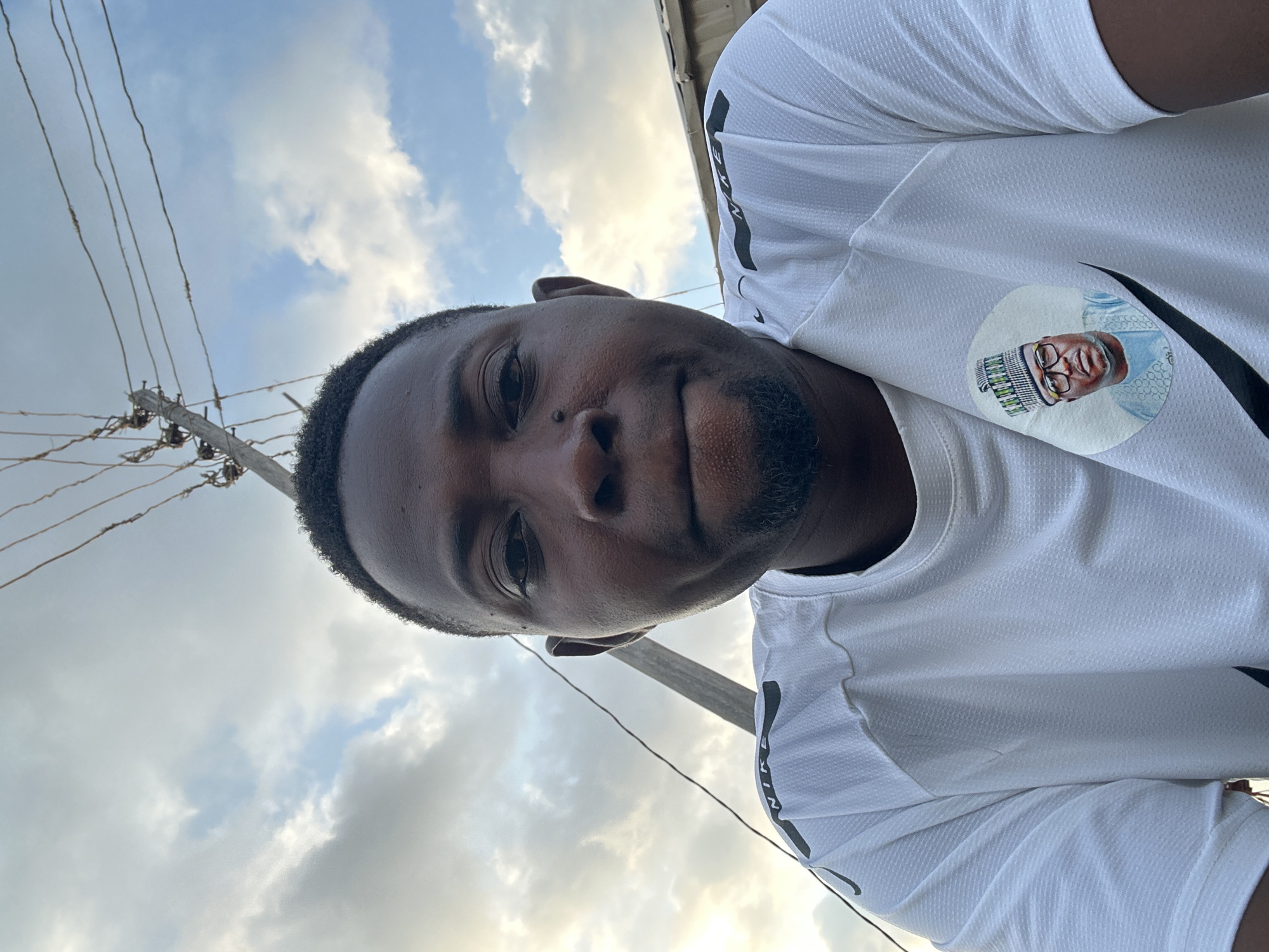 A close-up, low-angle outdoor selfie captures a man in Narayi, Nigeria, under a cloudy sky. He wears a white Nike t-shirt with a circular badge on the left side featuring a portrait of a man in traditional attire and glasses. The man in the selfie has dark skin, a short beard, and is looking directly at the camera. A wooden utility pole with wires runs diagonally through the background, with a portion of a corrugated metal roof visible at the bottom right.