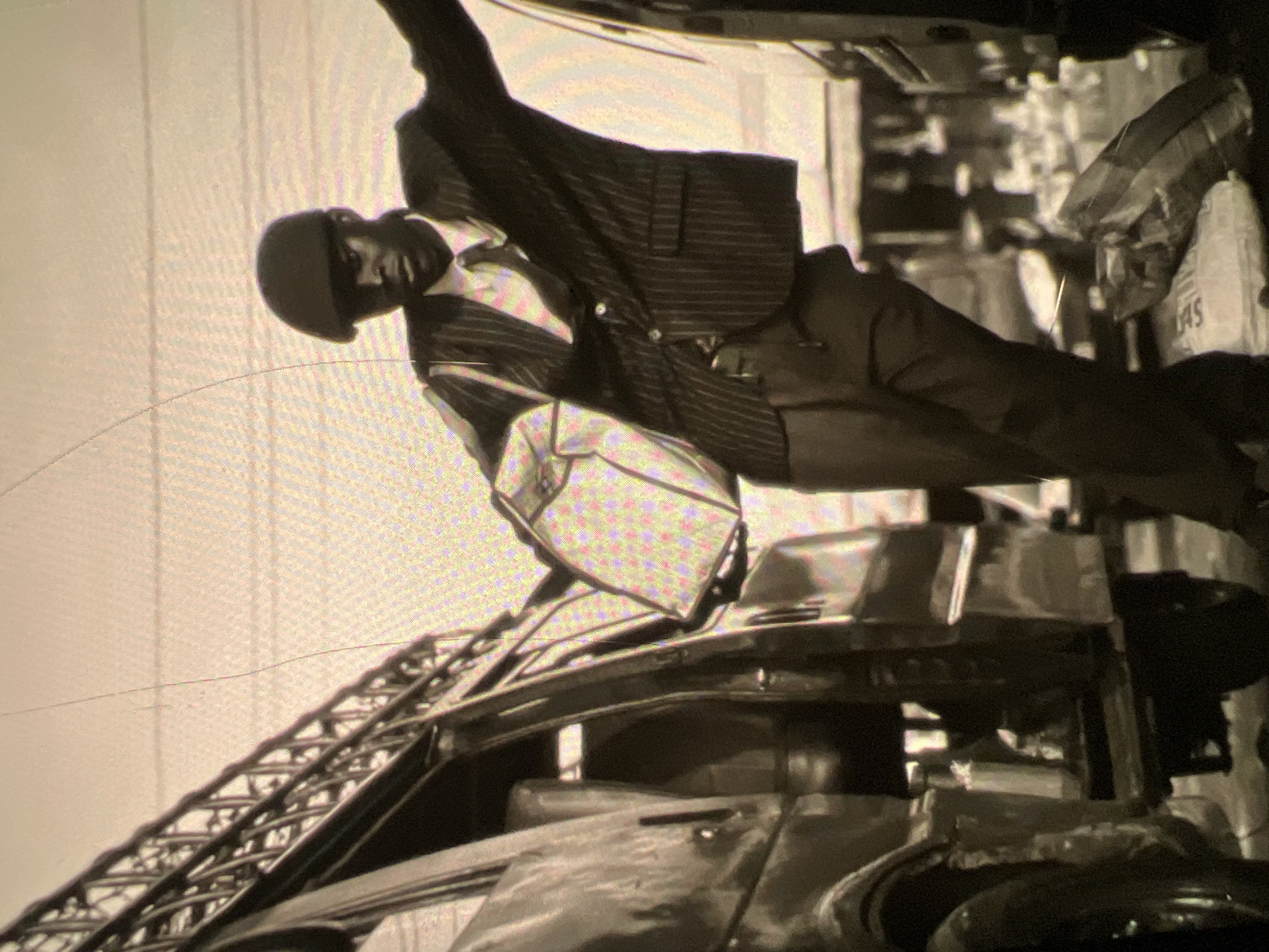 The black and white, slightly grainy photograph captures a scene on a bustling street in Lagos, Nigeria. A man, dressed in a pinstripe suit, white shirt, and tie, wears a flat cap and leans casually against an open vehicle. He is holding a large bag over his shoulder. The vehicle appears to be a utility van or a bus, with a metal structure, possibly a ladder or scaffolding, extending upwards from its side. In the background, there are more vehicles, suggesting a commercial area or a transport hub. The ground is littered with several large bags, some appearing to be sacks of goods. The lighting suggests it is daytime, and the overall impression is one of activity and commerce within the urban environment of Lagos.