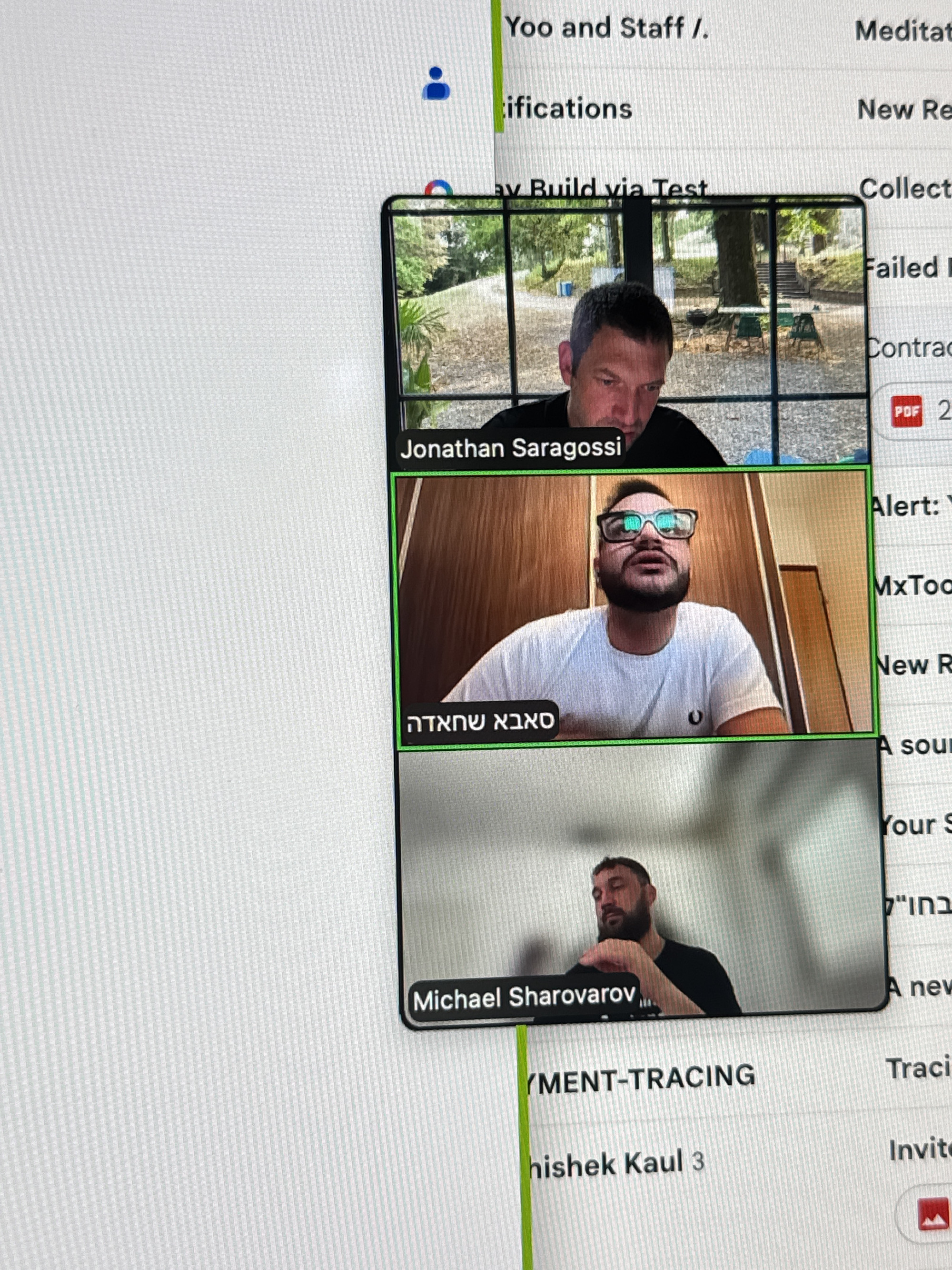 The image displays a computer screen with an open application, likely a video conferencing tool, showing multiple participants. The application is overlaid on what appears to be a desktop or web browser interface.
In the video conferencing window, there are three visible participants:
1. The top participant, identified by the name "Jonathan Saragossi," is a man with short dark hair, wearing a black t-shirt. He is looking downwards. The background of his video feed shows an outdoor scene with trees and stairs.
2. The middle participant is wearing glasses and a white t-shirt with a logo on the left chest. His name is displayed in Hebrew text: "סאבא שמחזיה" (Sabah Shamhaziya).
3. The bottom participant, identified as "Michael Sharovarov," is a man with a beard, wearing a dark t-shirt, and looking downwards. The background is a plain wall.
To the right of the video conferencing window, there are text entries that appear to be from the surrounding application or operating system. These include "Yoo and Staff /.", "Meditat", "ifications", "New Re", "av Build via Test", "Collect", "Failed R", "Contrac", and "Alert: Y", "MxToo", "New R", "A sour", "Your S", and text in Hebrew that appears to read "בחו"י" (Bchuyi), followed by "A new". Further down, text includes "YMENT-TRACING", "Traci", "hishek Kaul 3", and "Invite". There is also a small red icon that resembles a mountain, possibly indicating a file or attachment.
The image is a close-up of a computer screen, showing the typical pixelated texture of a digital display. There is no indication of the city of Erba, Italy, within the visible content of the image. The setting appears to be indoors, in front of a computer.