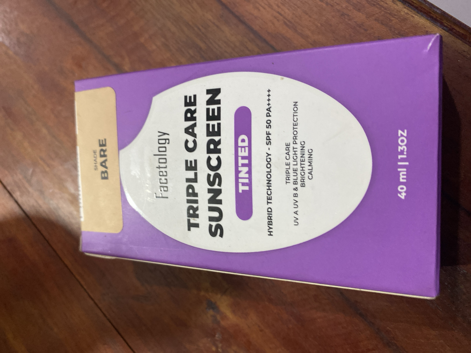 A close-up, slightly angled shot focuses on the front of a purple and tan cardboard box for "Facetology Triple Care Sunscreen" with the shade "Bare" and the text "Tinted" in a purple oval. The box displays product details such as "Hybrid Technology - SPF 50 PA++++" and lists benefits like "Triple Care," "UVA UVB & Blue Light Protection," "Brightening," and "Calming." Below this, the volume "40 ml | 1.30Z" is visible. The box rests on a wooden surface with a visible grain pattern. The image was taken in Ta', Indonesia.