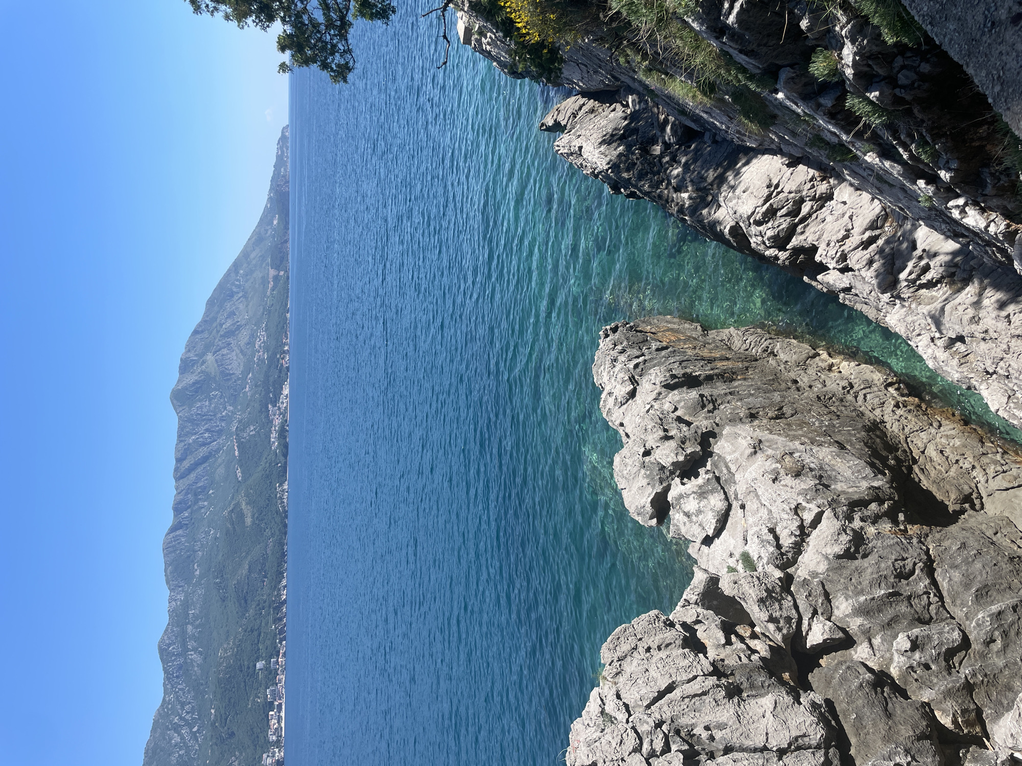 The image captures a scene from Bečići, Montenegro, featuring a steep, rocky coastline overlooking the Adriatic Sea. The water is clear, with a gradient of turquoise near the rocks transitioning to deeper blue further out. The rocky cliffs are composed of light gray, textured stone, with some vegetation growing on the edges. In the distance, a mountain range stretches along the coastline, with signs of habitation at its base. The sky is a clear, bright blue. The overall impression is one of a rugged, natural landscape.