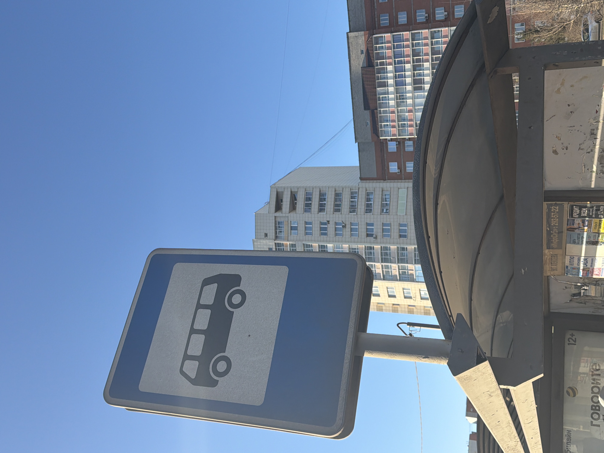 The image shows a bus stop in Krasnoyarsk, Russia. The stop is marked by a blue and white sign depicting a bus. A metal shelter provides a small space for waiting passengers, with advertisements and notices posted on its inner surfaces. Tall buildings can be seen in the background, indicating an urban setting. The sky is a clear blue.
