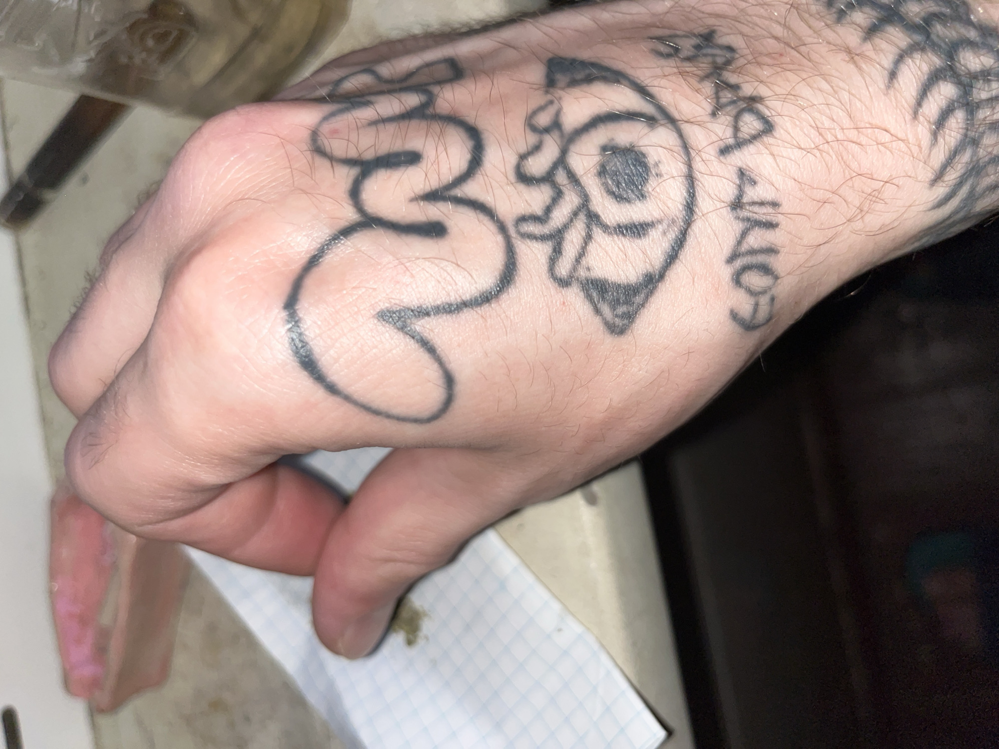 A close-up shot shows a person's hand, tattooed with black ink, resting on a surface that appears to be a countertop. The tattoos include what looks like abstract lettering on the back of the hand, an eye with what seems to be a hand or wing underneath it, and more abstract designs on the wrist and side of the hand. A small piece of squared paper with some green material on it is visible on the left side of the frame. In the background, out of focus, are what appear to be a pink object, a glass container with a brownish liquid, and a dark object that could be a cigarette or blunt. The lighting suggests an indoor environment.
