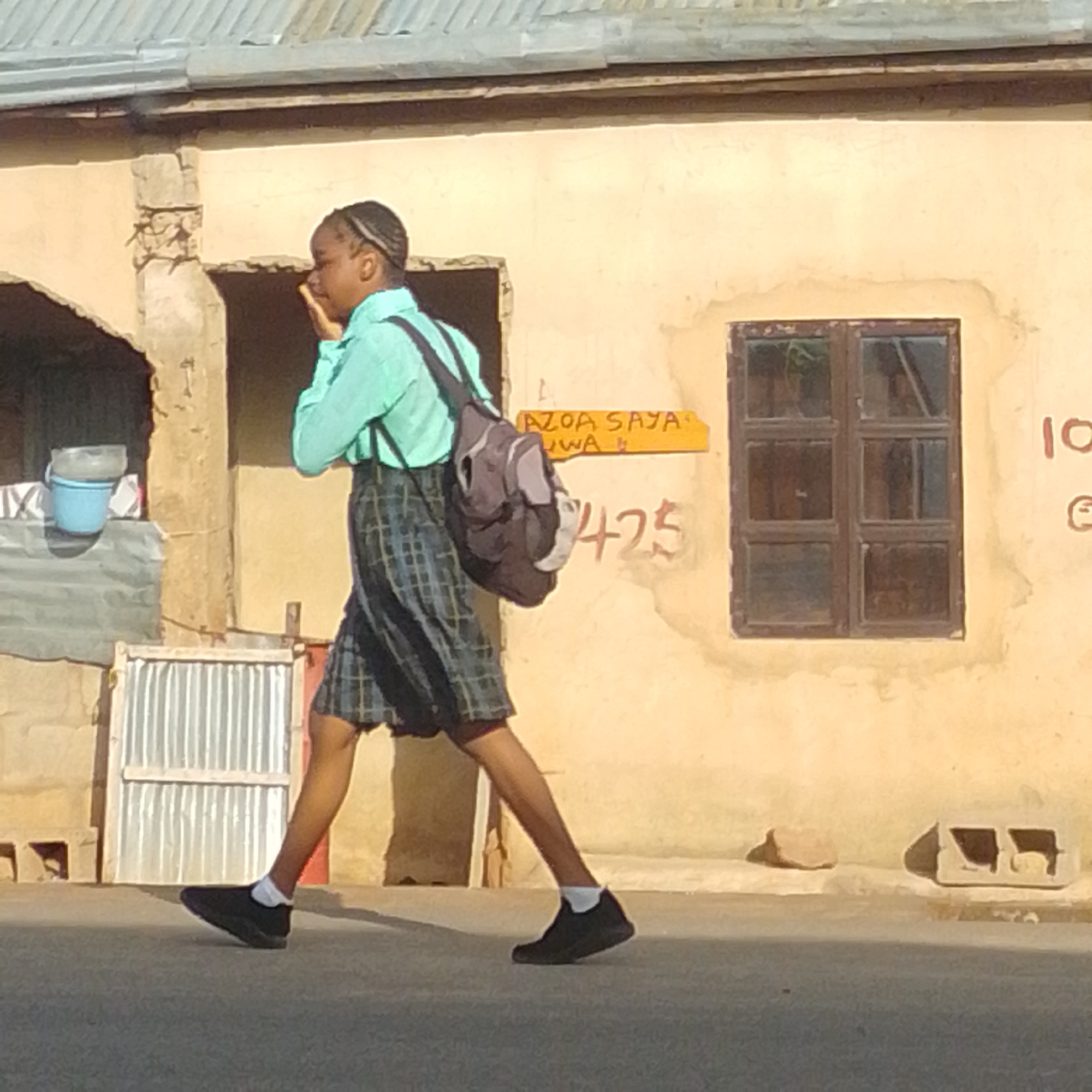 A young person, likely a student, walks along a paved street in front of a tan-colored building. The individual is wearing a light blue collared shirt and a plaid skirt, along with black shoes and white socks. A dark gray backpack is slung over their left shoulder. The building features a corrugated metal roof and a wooden-framed window with multiple panes. A yellow sign with orange lettering is affixed to the wall, displaying the text "AZOA SAYA" and "JUWA" followed by a number that appears to be "425". To the left of the building, there is an open doorway with a blue bucket and a clear plastic container visible inside. Further left, a section of corrugated metal is attached to a white frame. The ground is a flat, dark gray surface, and a few concrete blocks and stones are scattered on the edge of the pavement. The lighting suggests it is daytime, with shadows cast on the ground. The image is taken from a slightly low angle.
