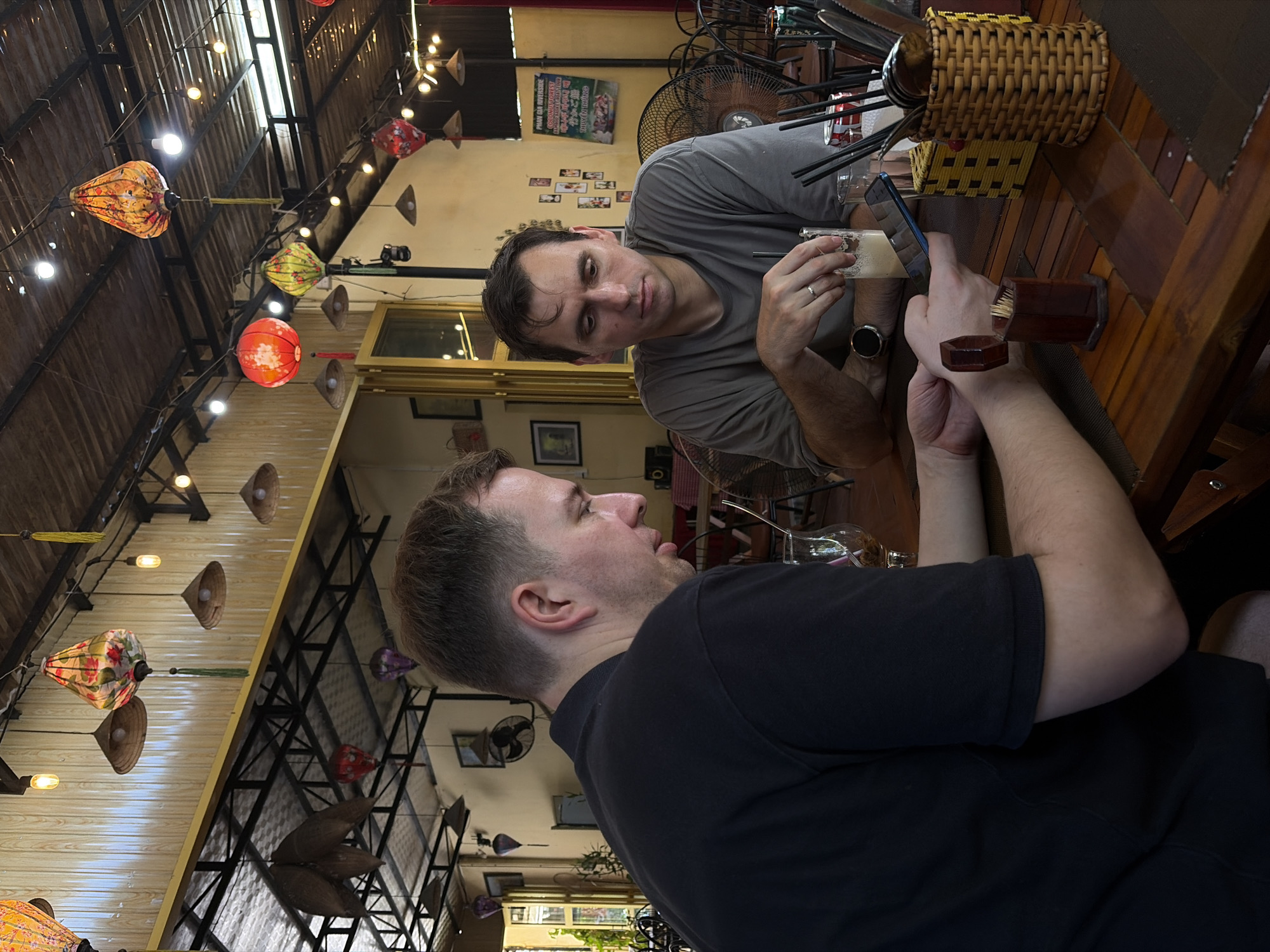 Two men are seated at a wooden table in what appears to be a lively Vietnamese restaurant or cafe, consistent with the provided location context of Đà Nẵng. The original image is rotated, showing the scene from a sideways perspective.
The man on the right, wearing a grey T-shirt, is looking towards the man opposite him. He holds a glass with a light-colored beverage, possibly a beer or frothy drink, in his left hand, and has a watch on his left wrist. A wedding band is visible on his left ring finger. A smartphone is also on the table near his right hand.
The man on the left, wearing a black T-shirt, is facing the man in grey, with his tongue playfully extended. His expression suggests a lighthearted or humorous interaction.
The table between them holds a woven rattan basket filled with utensils (chopsticks, spoons) and a wooden holder with toothpicks.
The background features a ceiling decorated with numerous colorful lanterns, typical of Vietnamese craftsmanship (e.g., Hoi An style lanterns common in the region). Interspersed among the lanterns are traditional Vietnamese conical hats (Nón Lá) used as decorative elements and strings of small light bulbs. The ceiling appears to be made of wood planks and exposed metal beams. Several fans are visible in the background, suggesting a warm climate. On a yellow wall in the background, there are framed pictures and a prominent green sign with Vietnamese text, reading "PHỞ GÀ ĐẶC BIỆT," which translates to "Special Chicken Pho," further confirming the Vietnamese setting. Sunlight or bright natural light is visible through windows or open sections of the establishment.