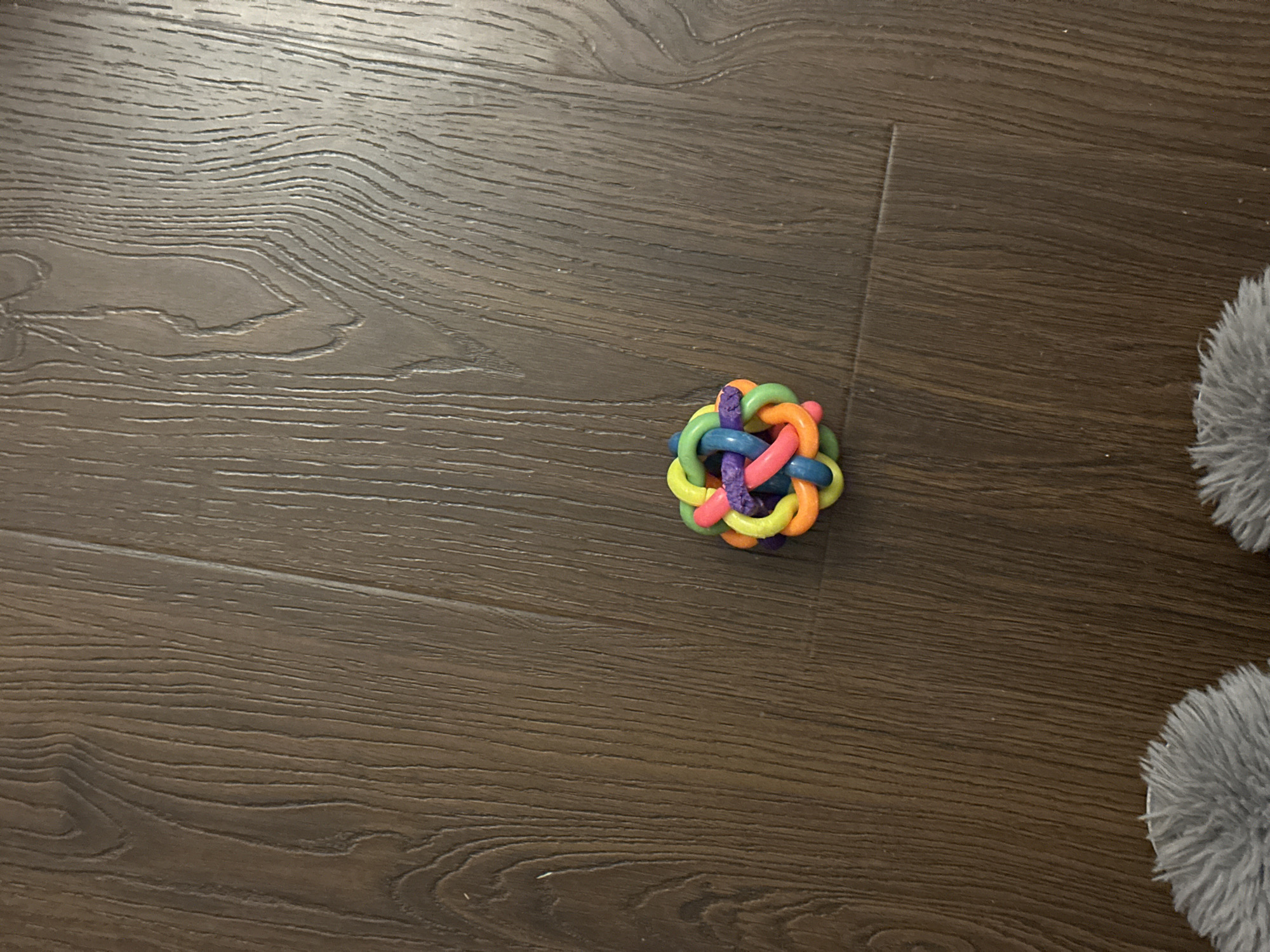 A colorful, woven ball toy rests on a dark wooden floor. The toy is approximately spherical and composed of intertwined plastic loops in various bright colors, including yellow, green, orange, blue, and pink, with a purple core. The floorboards are dark brown with visible wood grain patterns running diagonally across the image. In the lower portion of the frame, the edge of two fluffy, grey slippers is visible, suggesting someone is wearing them. The scene is indoors, with no indication of a specific city or location like Verkhniy Yurt, Russia.