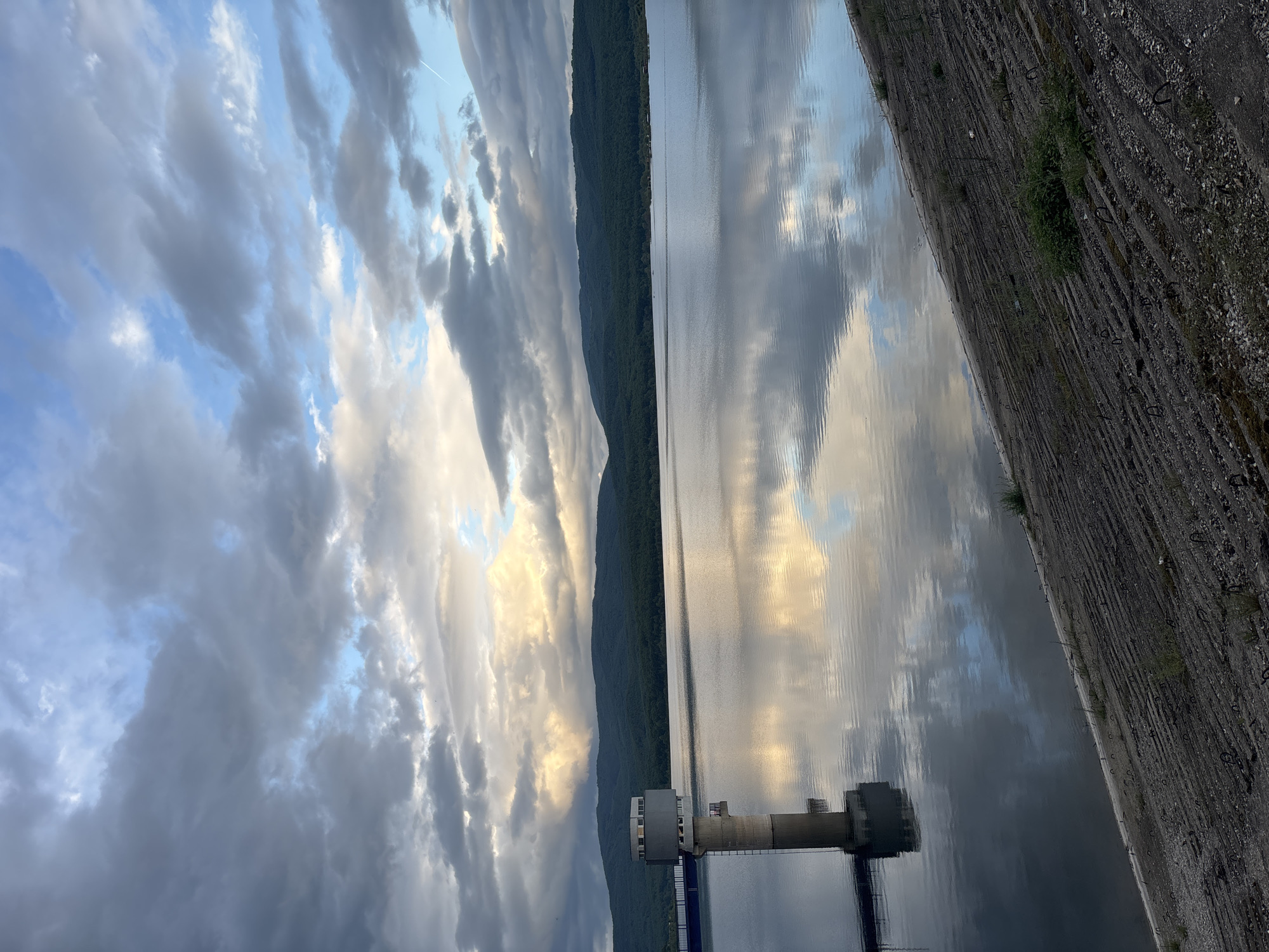 The image is a horizontal view of Ognyanovo, Bulgaria, featuring a reservoir with a concrete dam on the right. In the lower part of the photo, there's a tower structure extending into the water. Across the reservoir, there's a forest, with a view of a dramatic cloudy sky in the background. The surface of the water mirrors the sky and surroundings.