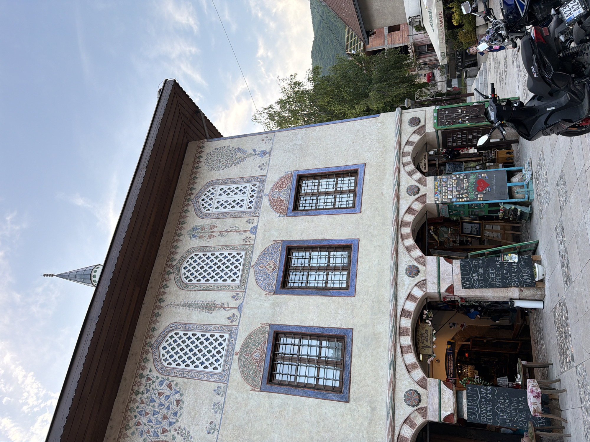 This is a photograph of the Colored Mosque in Travnik, Bosnia and Herzegovina. The mosque is a historical building known for its colorful exterior decorations. In the foreground, there are several motorcycles parked on a cobblestone street. The building itself features arched entrances on the ground floor, with shops or stalls set up behind them. The upper floors have distinctive windows and intricate decorative patterns. A young girl is visible in the background, walking on a street that leads to other buildings and a shaded area with seating. The sky is partly cloudy, and a green hillside is visible behind the buildings.