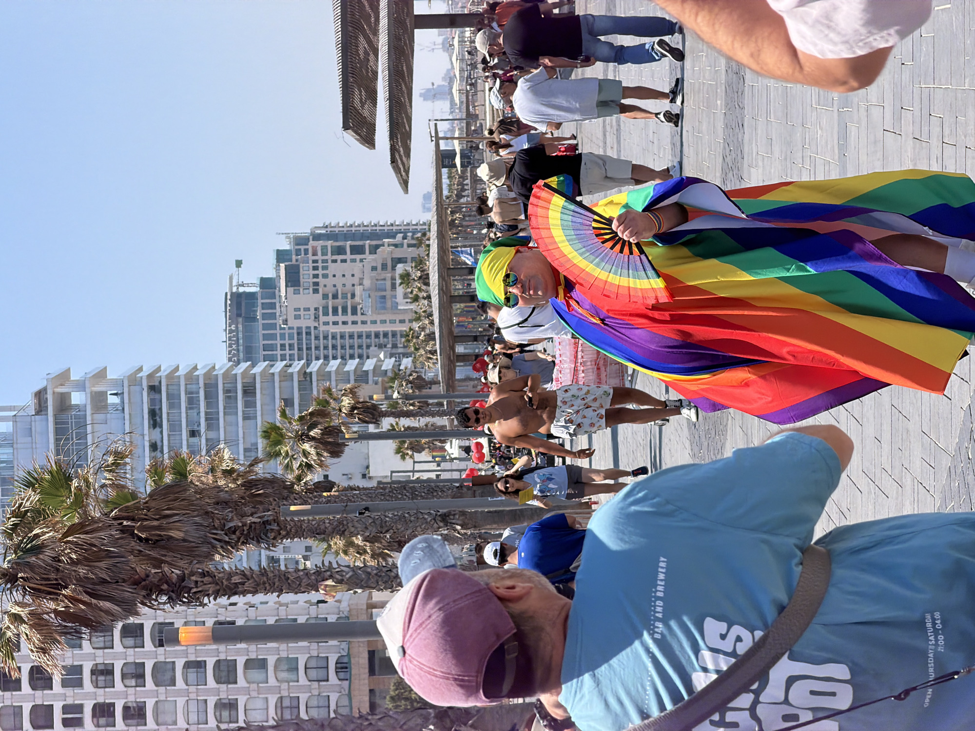 The image depicts a street scene in Tel Aviv, Israel, likely during an outdoor event or festival. In the foreground, two individuals are partially visible. The person on the left, seen from behind, wears a light blue t-shirt with text that includes "BAR AND BREWERY". They are also wearing a maroon baseball cap. To their right, another person is prominently featured, dressed in a vibrant rainbow-colored garment, possibly a cape or robe, adorned with a rainbow fan in their hand. They are also wearing a green headscarf and sunglasses.
In the background, a crowd of people are walking along a paved walkway lined with palm trees. Tall, modern buildings with many windows are visible behind the trees, characteristic of Tel Aviv's urban landscape. The sky is clear and blue, indicating a sunny day. There are also some structures providing shade, suggesting a public area or promenade. The overall atmosphere appears to be festive and lively, with many individuals dressed in casual summer attire. The presence of rainbow colors strongly suggests a Pride event.