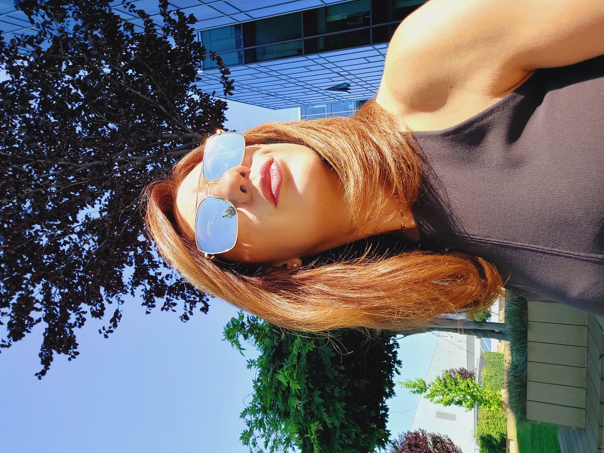 A medium shot photograph captures a woman from the shoulders up, looking to her right and slightly upward. She has auburn hair, styled to shoulder length, and is wearing reflective aviator sunglasses. Her lips are slightly parted, and she appears to be in an outdoor urban setting. To her right, a modern glass-fronted building stands tall, its facade reflecting the blue sky and trees. In the background, a dark-leaved tree branches out over her, and to her left, a well-manicured green tree and manicured bushes are visible. The scene is bathed in bright sunlight, casting shadows and highlighting the woman's arm and face.