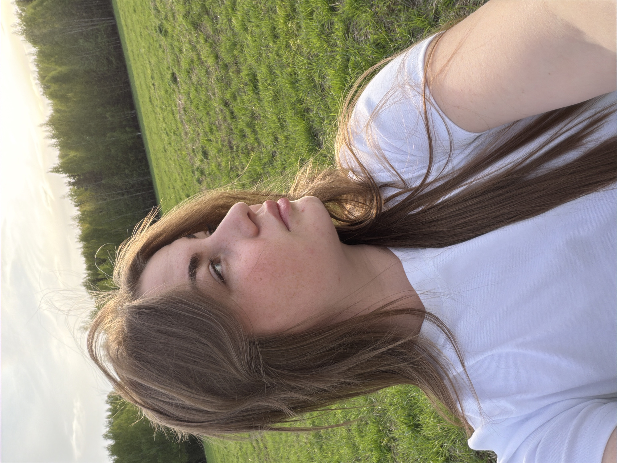 A young woman with light brown hair and fair skin is lying on her back in a grassy field. She is wearing a white t-shirt. Her head is turned slightly to the left, and she is looking upwards. The field is green and appears to be a meadow or pasture. In the background, there is a line of trees, suggesting a forest or wooded area. The lighting suggests it is late afternoon or early evening. The image is not related to Russia or the city of Zlodar'.
