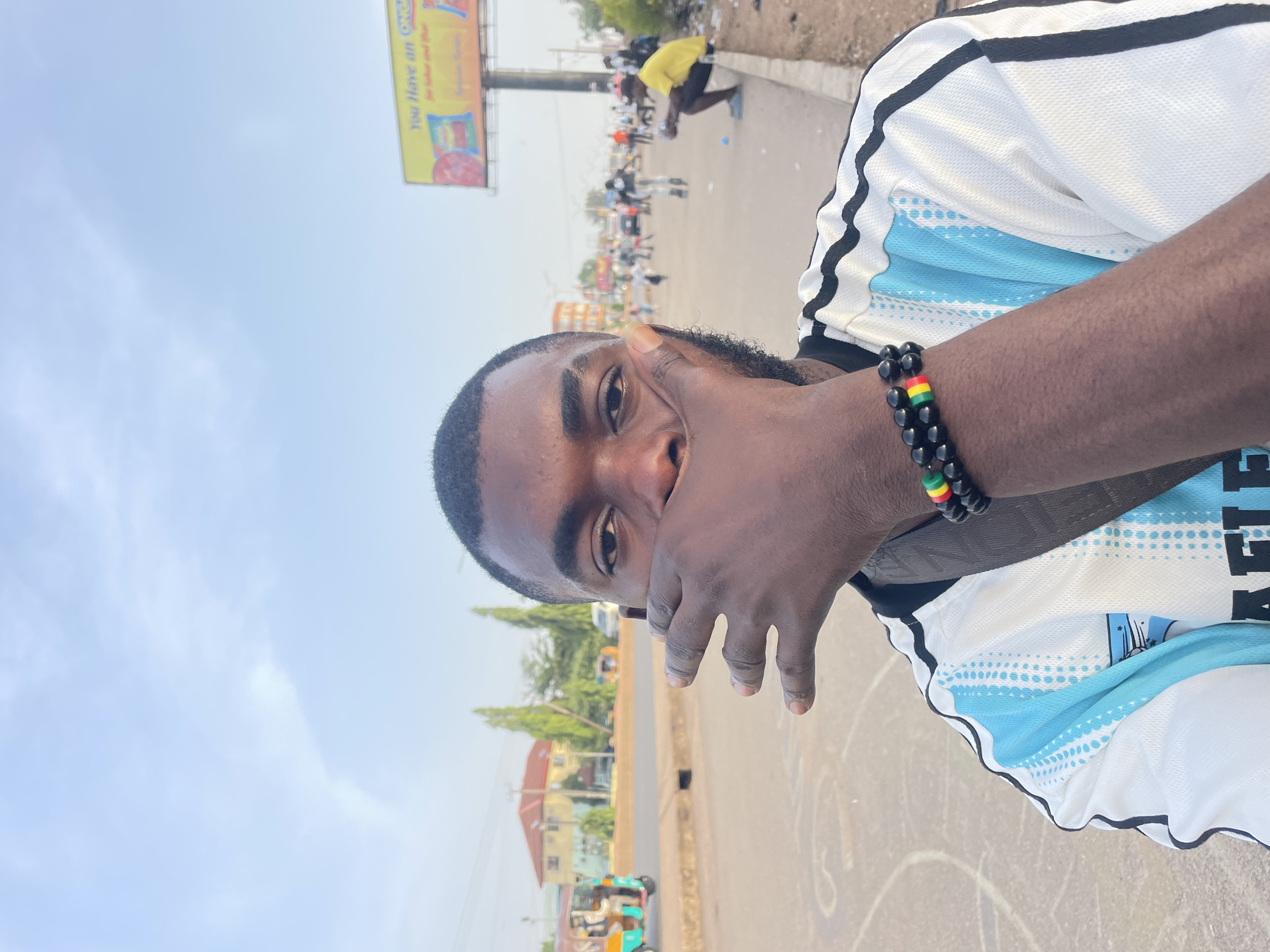 A portrait-oriented photograph captures a young Black man from the chest up, looking directly at the camera with a slight smile. He has short, dark hair, a beard, and his left hand is partially covering his mouth and chin. He is wearing a white sports jersey with light blue patterned accents and black stripes on the shoulders. On his right wrist, he wears two beaded bracelets: one is solid black, and the other features black, red, yellow, and green beads.
The background reveals an outdoor urban scene under a bright, clear blue sky, suggesting daytime. A wide, paved road stretches into the distance, with people walking and standing along its sides. One individual in a bright yellow top is visible bending over near the roadside in the mid-ground. Further back, buildings with orange-brown roofs and green trees are discernible. A colorful motorized tricycle, likely a "keke napep" common in Nigeria, is partially visible on the left side of the road in the distance. On the right side, a large billboard mounted on a pole shows the partial text "You Have an ON..." and "for labour and time". The overall setting depicts a lively street in Jos, Nigeria.