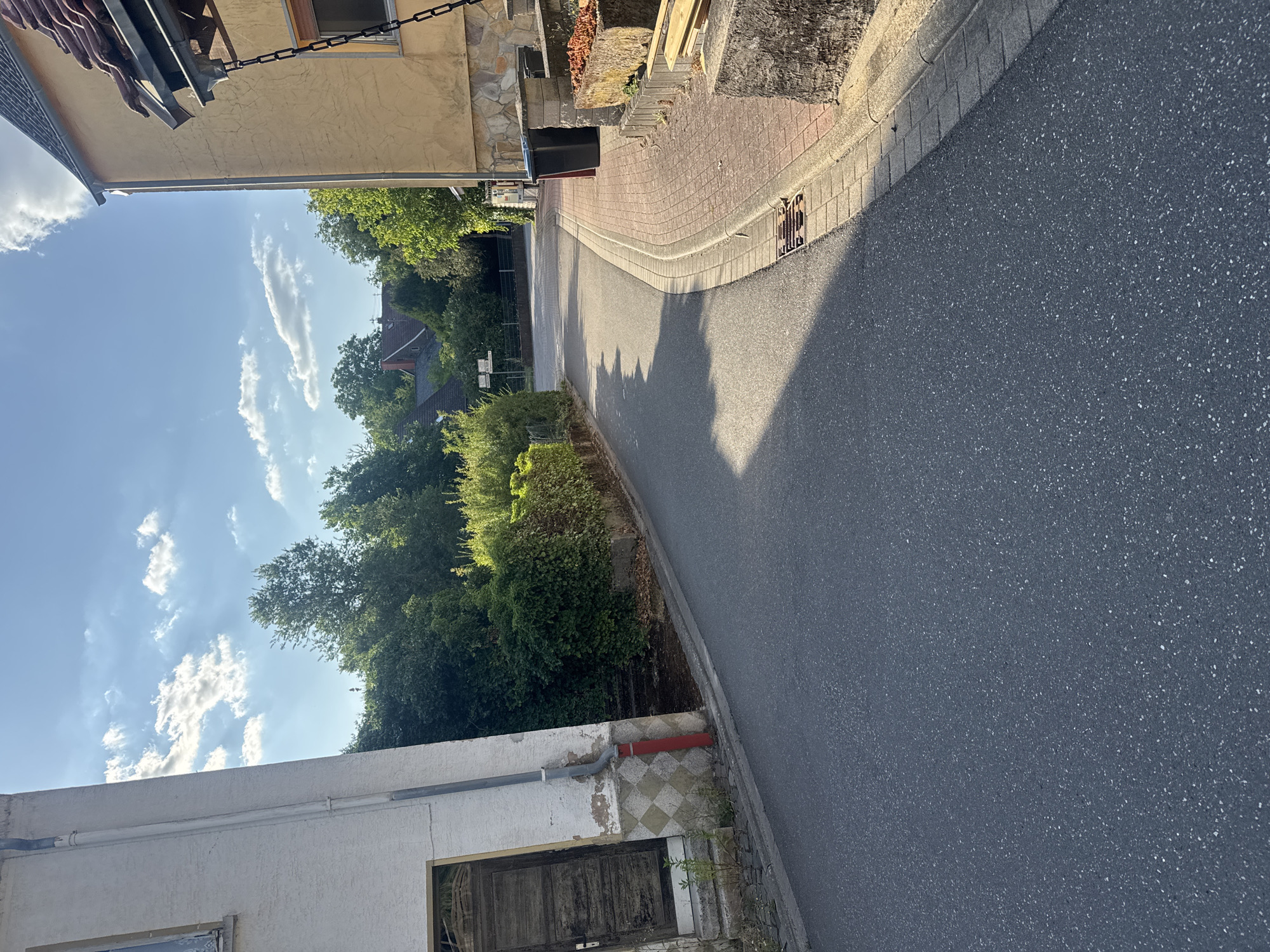 The image depicts a street scene in Hüffler, Germany, on a sunny day with scattered clouds. The perspective is from street level, looking down a paved road that curves to the right.
On the left side of the street, a white building with a weathered wooden door is partially visible. A gray downspout runs along the side of this building, with a red pipe section near the bottom. The building has a textured facade and a checkered tile pattern at its base.
The right side of the street features a yellow building with a tiled roof and a stone-clad lower section. Two dark-colored trash bins are situated near the yellow building. A paved walkway with red brick pavers runs alongside the road. A concrete planter box filled with dry vegetation is also present.
Mature green trees and dense hedges line both sides of the street, creating a suburban or village atmosphere. In the background, several houses with red-tiled roofs are visible, nestled among the trees. Road signs are faintly discernible in the distance. The road surface is asphalt, and a storm drain grate is visible on the right side. The sunlight casts long shadows across the road.