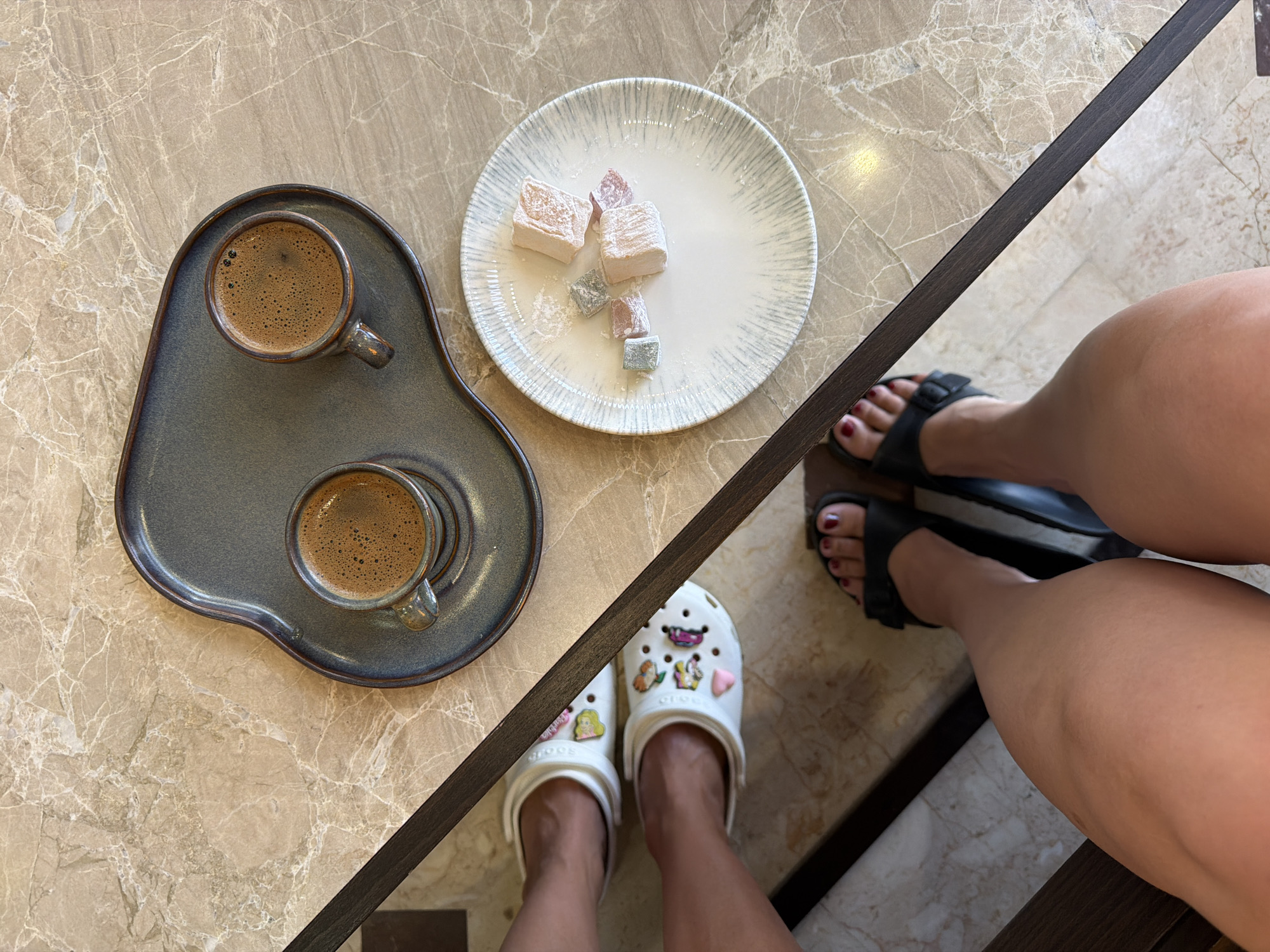 An overhead shot shows two cups of Turkish coffee on a dark gray serving tray, positioned on a marble table. To the left of the tray are two white Crocs shoes adorned with various colorful charms. To the right of the tray is a white plate with a patterned rim, holding several pieces of Turkish Delight. In the foreground, the lower legs and feet of two individuals are visible. One person wears the decorated Crocs, and the other wears black sandals. The setting appears to be a cafe or restaurant in Manavgat, Turkey, given the presence of Turkish coffee and Turkish Delight.