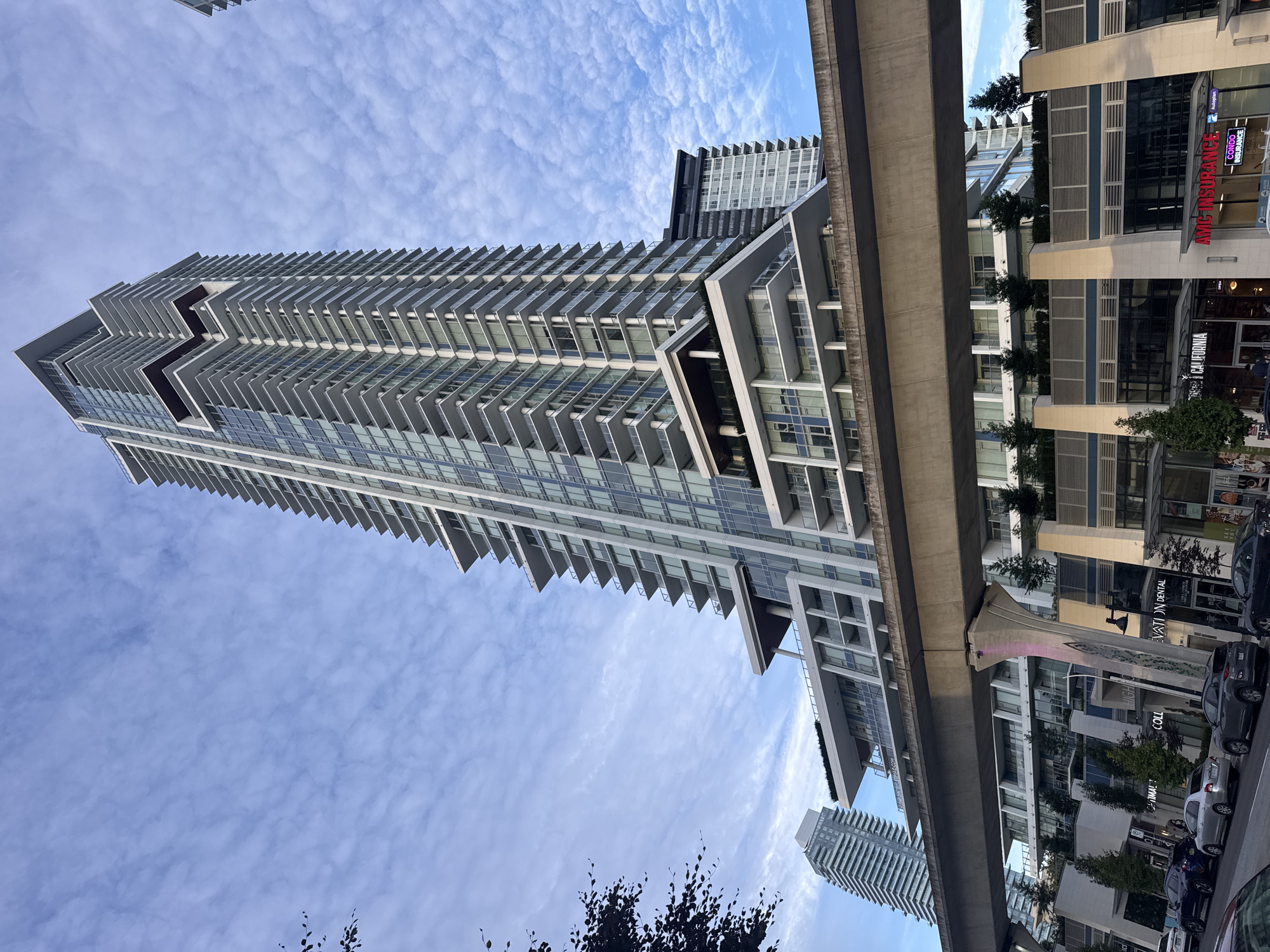 A low-angle, wide shot captures a modern skyscraper dominating the frame under a cloudy, blue sky. The skyscraper, with its sleek, glass facade and numerous balconies, is the primary subject. In the foreground, a concrete elevated structure, likely a transit line, cuts horizontally across the image, supported by angled concrete pillars. Below this structure, the ground level of the city is visible, featuring retail storefronts with signage such as "CALIFORNIA" and "AMC INSURANCE." Several cars are parked along the street. Another, shorter skyscraper is visible in the background to the left, and a portion of a third building appears to the right of the main skyscraper. The scene appears to be in a bustling urban environment, possibly in Coquitlam, Canada, given the context.