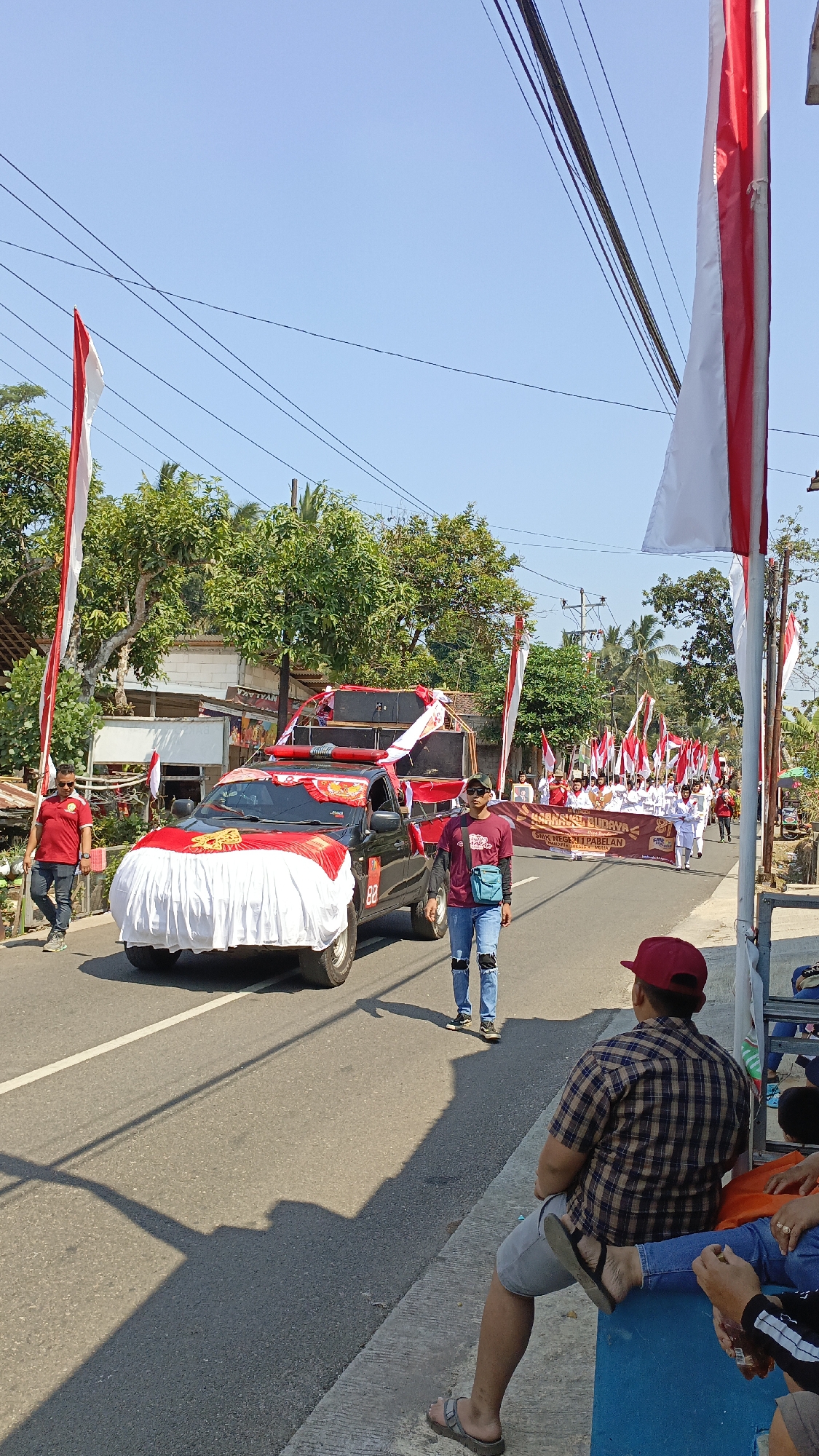 A daytime outdoor procession is underway on a paved street in Krajan Lor, Indonesia. The focus is on a black pickup truck decorated with red and white fabric and large speakers. The truck is draped with a white sheet at the front, adorned with red and gold embellishments. Two individuals, dressed in red shirts and jeans, are walking on the left side of the truck. Another person, wearing a maroon t-shirt and jeans, stands near the truck, looking towards the procession. In the background, a group of people in white outfits, carrying numerous Indonesian flags and a banner, are marching down the street. The scene is lined with trees and buildings. Several Indonesian flags are also displayed along the sides of the road. Spectators are visible sitting on the curb on the right side of the image. The sky is clear and blue.