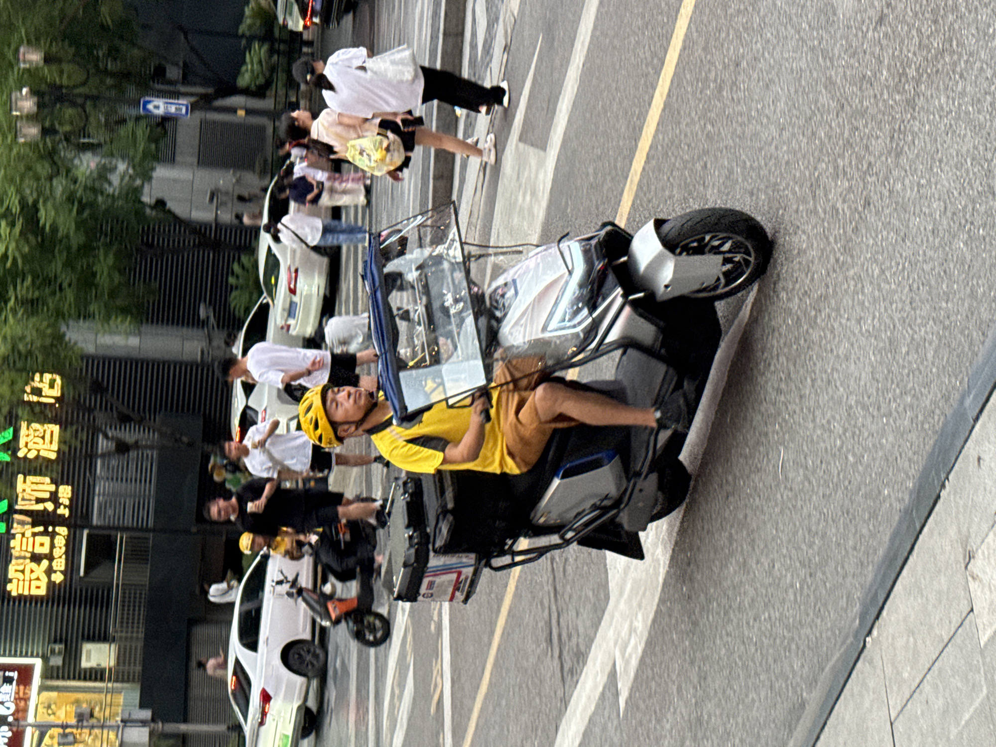 The image captures a street scene in Yuzhong District, China, focusing on a person riding a scooter. The rider, wearing a yellow helmet and a yellow t-shirt with brown shorts, is positioned in the foreground, operating a silver scooter with a large black storage box on the back. The scooter has a transparent shield in front of the rider.
In the background, several other individuals are visible, some walking and others standing. Two white cars are parked on the left side of the street. The environment appears to be an urban setting with buildings, trees, and streetlights. The street is paved with asphalt and marked with white and yellow lines, indicating a crosswalk. The overall impression is of a busy street with pedestrian and vehicular traffic.