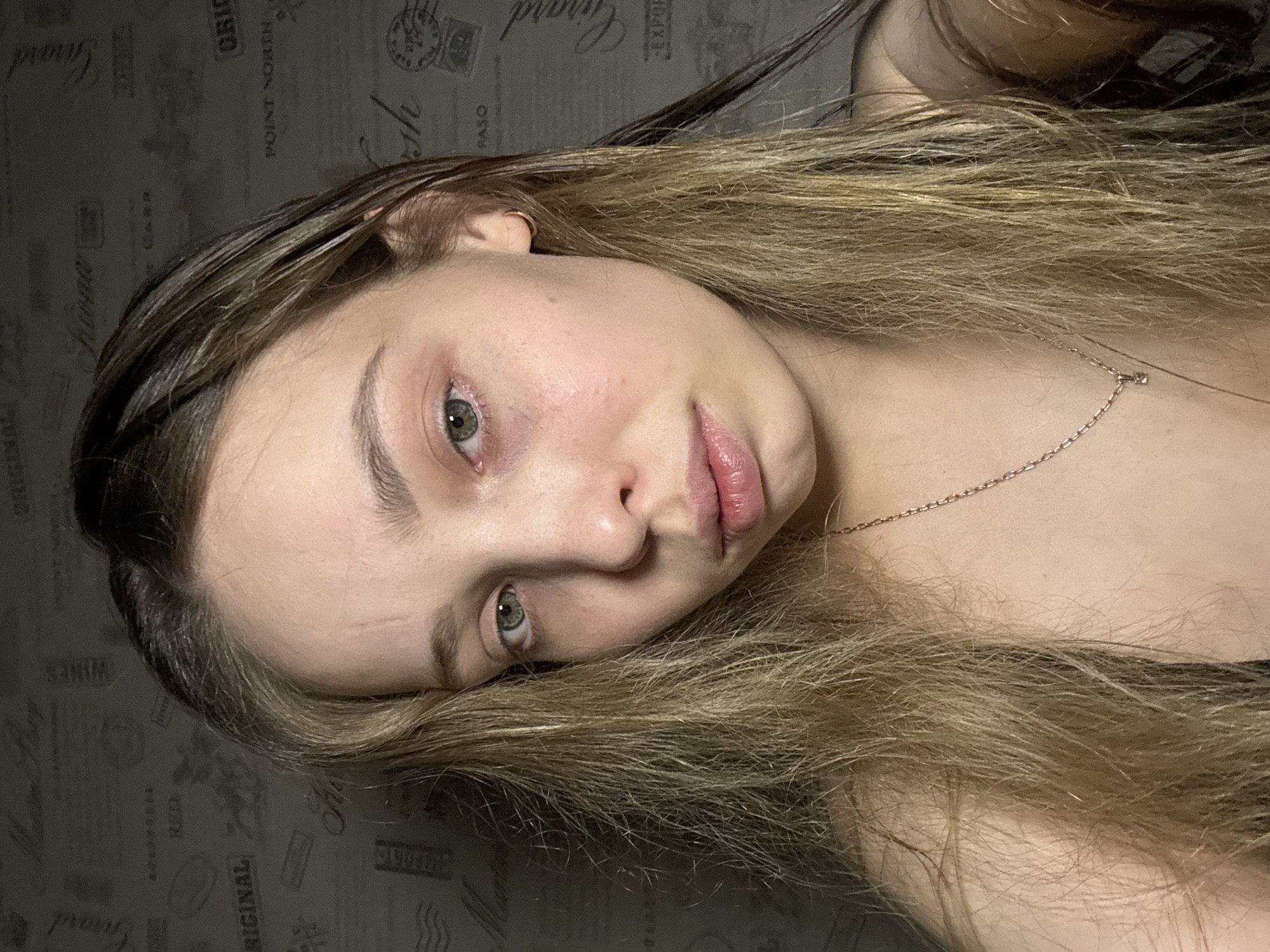 A close-up, slightly rotated portrait shows a woman with light brown hair, green eyes, and fair skin. The hair appears wet near the roots. She has a small gold hoop earring in her left ear and a delicate gold necklace. Her lips are full and slightly parted. The background appears to be a wall with a repeating pattern of vintage-style stamps or logos. The location of Chaikovsky, Russia is not evident in this image.