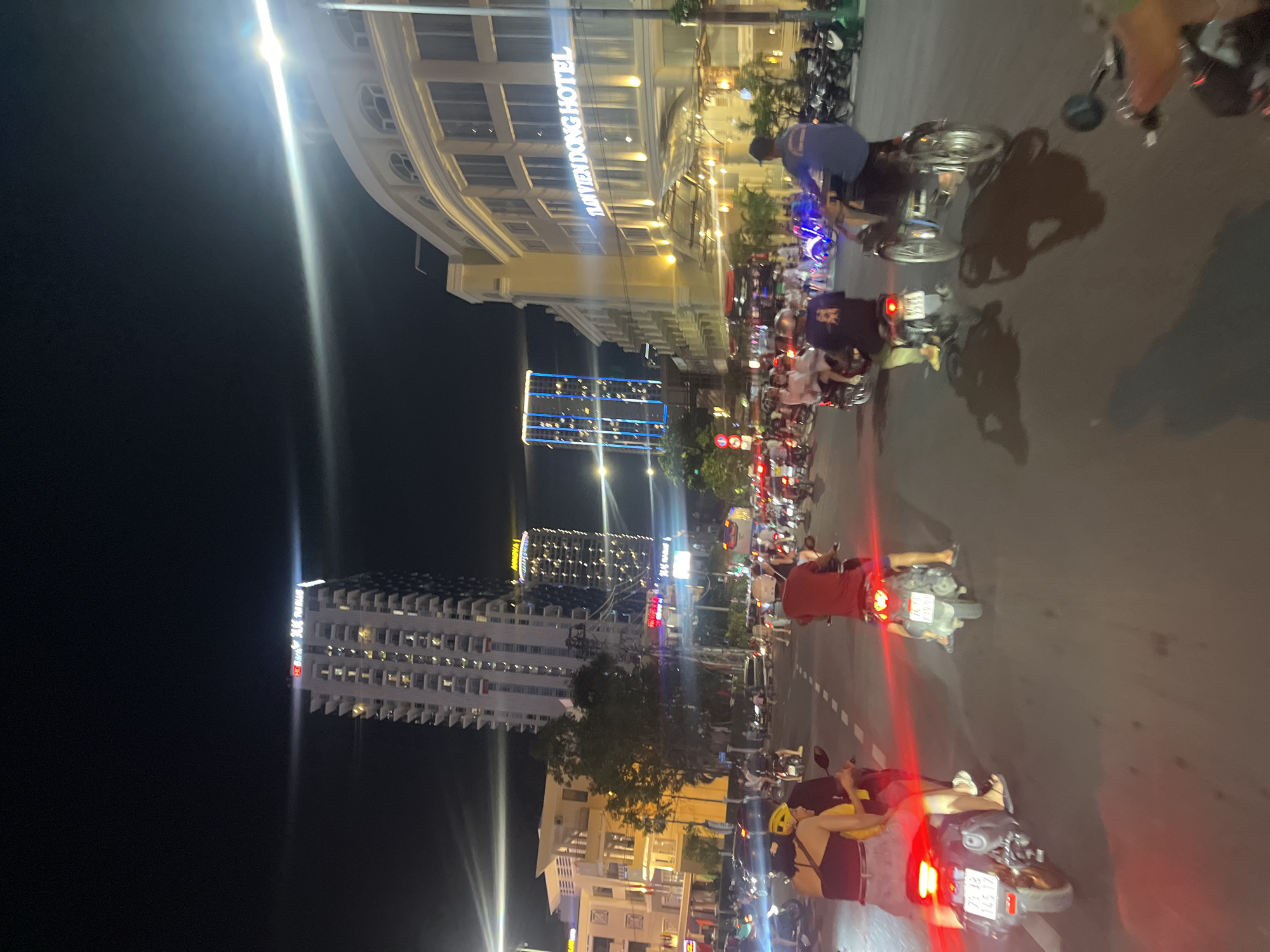 This is a nighttime street scene in Nha Trang Ward, Vietnam. A group of people are riding motorcycles in what appears to be a busy urban area. Buildings are visible in the background, including tall, modern structures and lower, more ornate buildings, some with illuminated signage. The road is paved and marked with white dashed lines. Streetlights and building lights illuminate the scene. Several motorcycles are the primary focus, with individuals riding them in various directions. Some motorcycles have their taillights illuminated, casting red glows on the road. The overall impression is of a lively, active street at night.