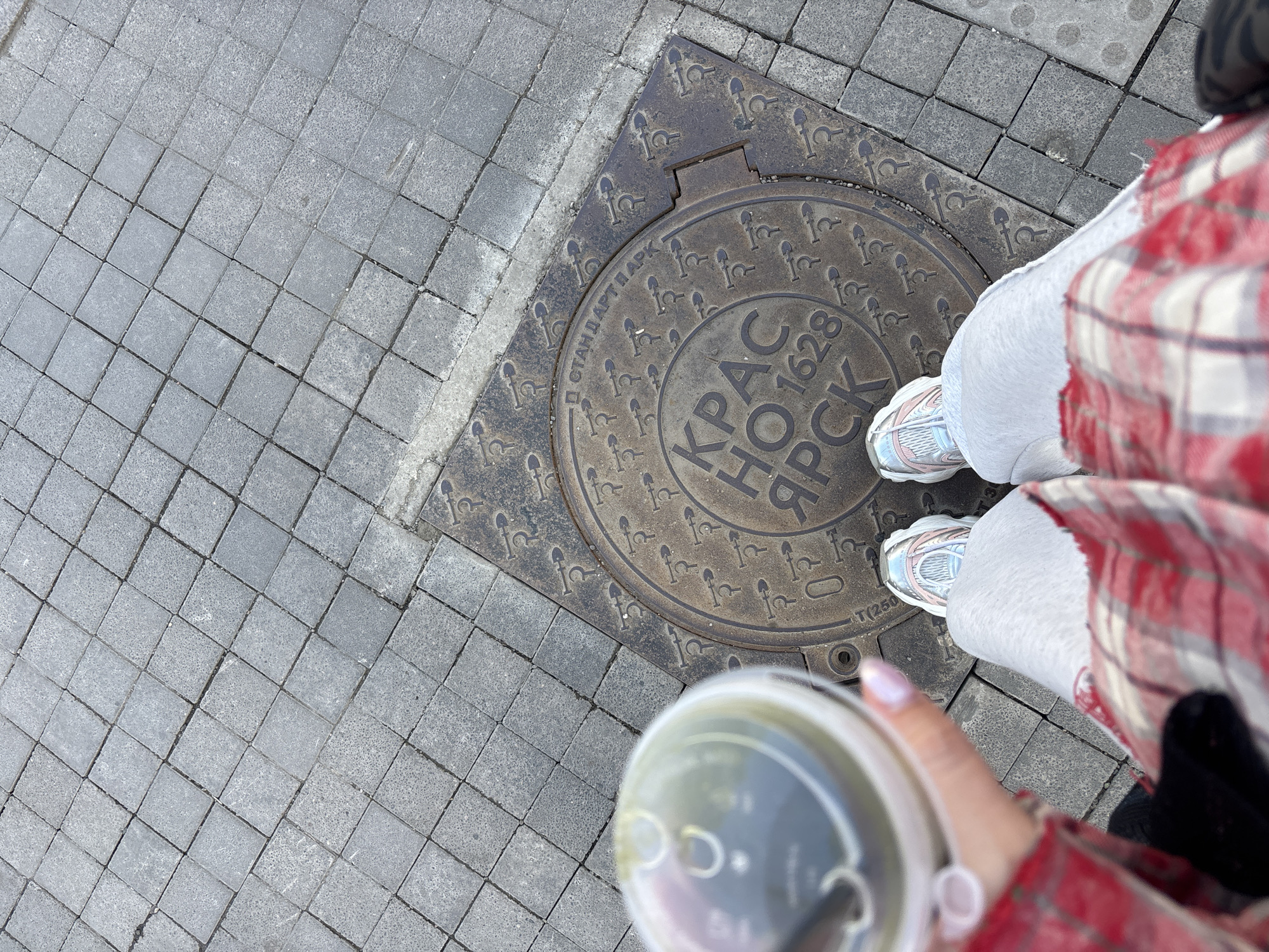 The image shows a person standing on a sidewalk in Krasnoyarsk, Russia. The person is wearing light-colored sweatpants, pink and white sneakers, and a red and white flannel shirt. They are holding a clear plastic cup with a lid and a straw. The person is standing on a manhole cover. The manhole cover is circular, and it has the words "Krasnoyarsk 1628" written in Cyrillic script. Around the text is a repeating image of a tool that looks like a shovel. The sidewalk is made of gray paving stones.
