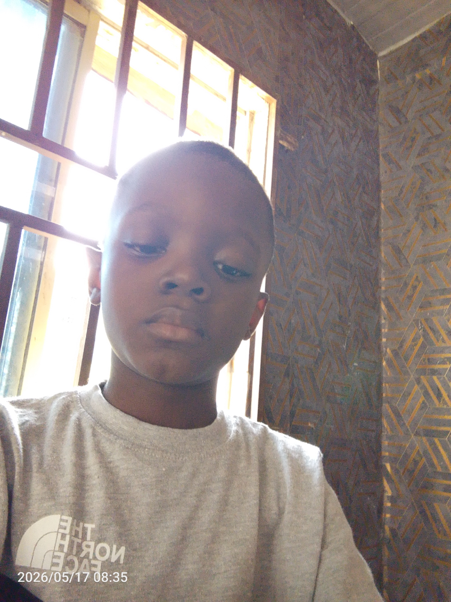 A young Black boy is indoors, standing near a window with bars in Lokoja, Nigeria. He is wearing a grey t-shirt with a white logo that appears to be reversed in the image. The wall behind him is decorated with a patterned wallpaper featuring geometric shapes. The timestamp "2026/05/17 08:35" is present on the bottom left of the image. The light coming from the window is bright, causing some glare on the boy's face.