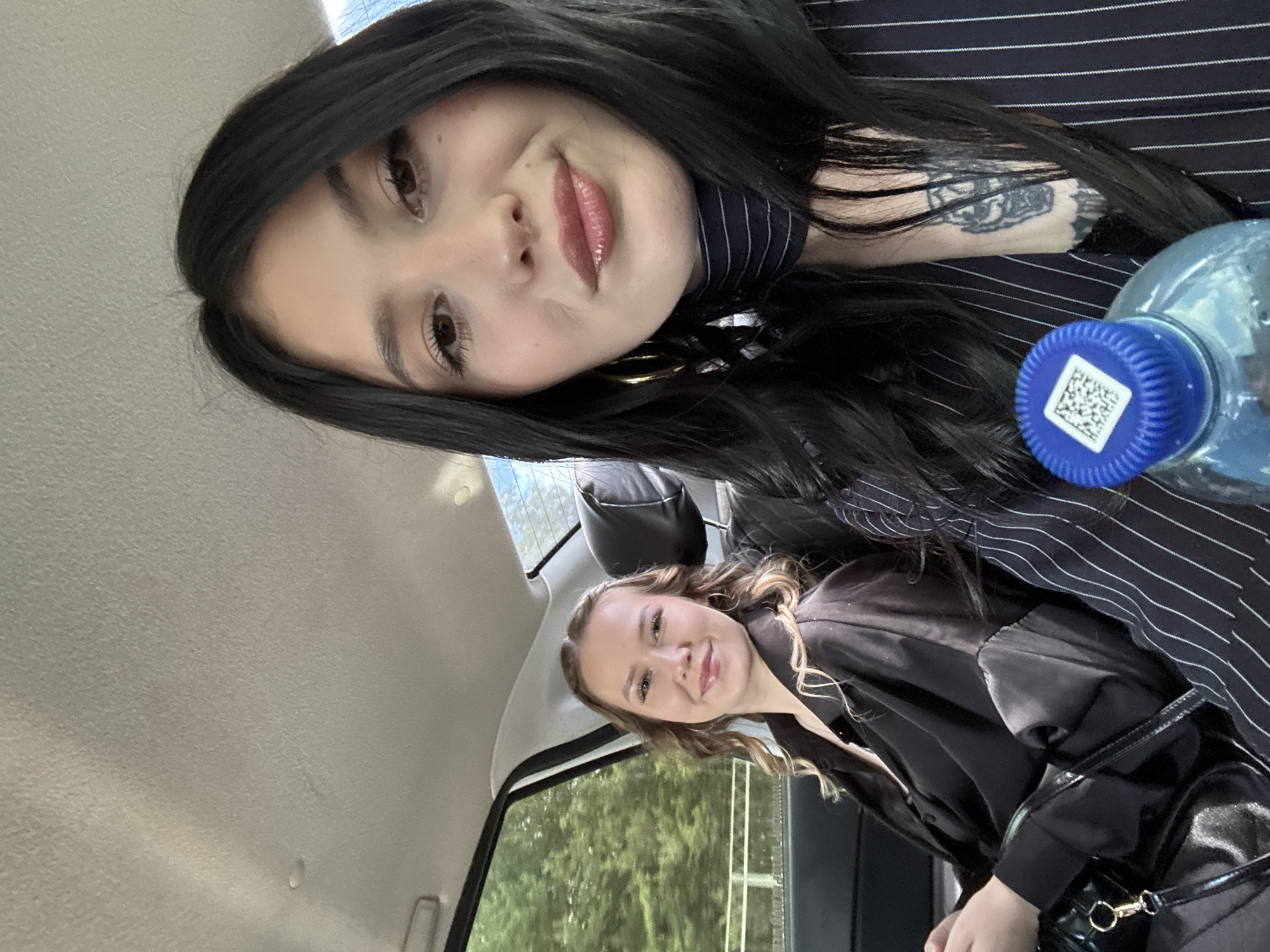 Two young women are seated in the back of a car, looking at the camera. The woman in the foreground on the right has long dark hair and is wearing a pinstriped jacket. She is holding a blue-capped bottle of water. The woman on the left has blonde hair and is wearing a black shirt. She is smiling. Outside the car window, trees and a fence are visible. The interior of the car, including the headliner and seats, is visible. The image was taken in the city of Chaikovsky, Russia.