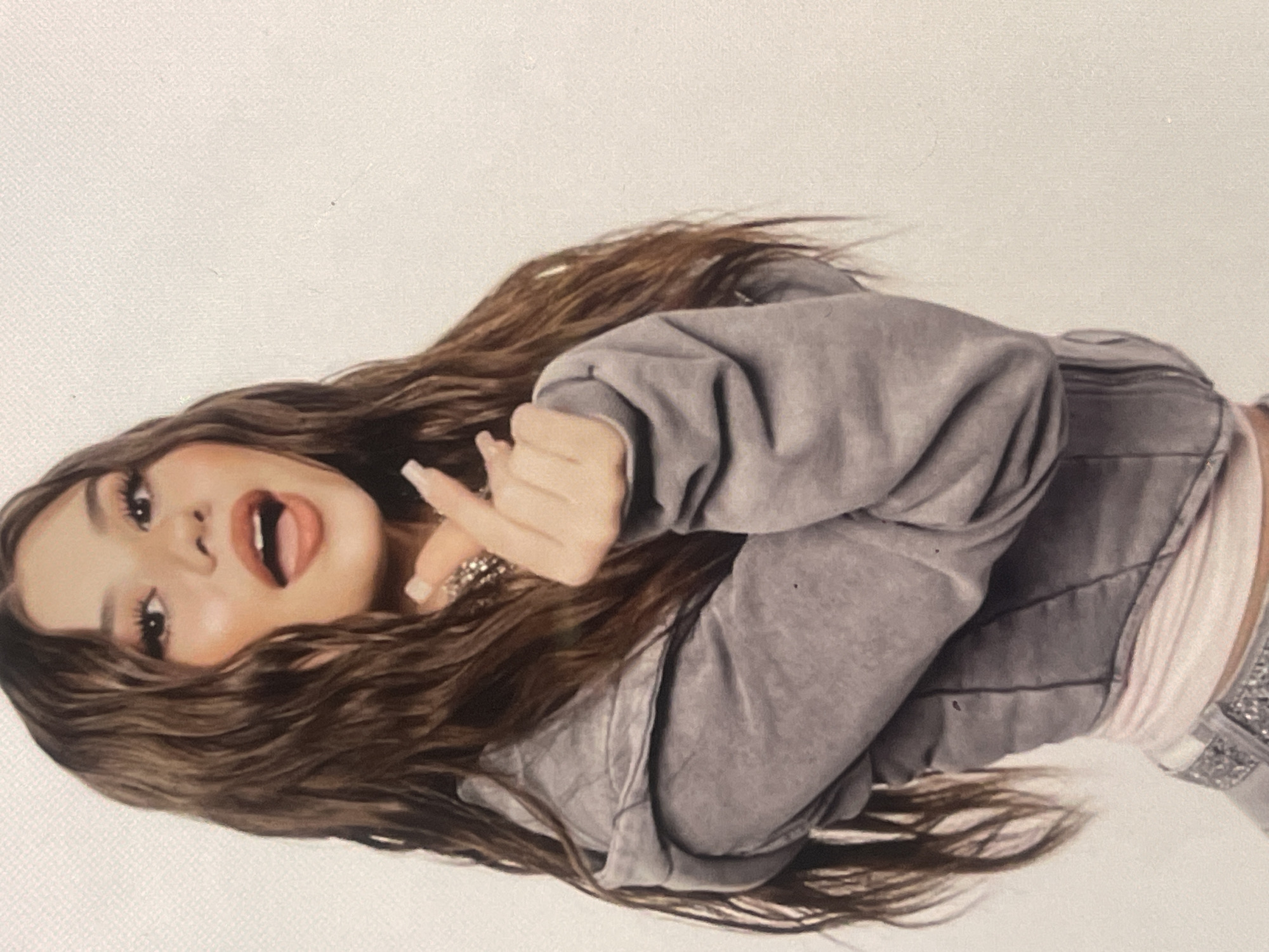 A young woman with long, wavy brown hair is depicted in a portrait-style image, rotated 90 degrees clockwise. She is positioned against a plain, light-colored background, possibly a studio wall or backdrop. Her face is heavily made up with prominent eyeliner, eyeshadow, and red-toned lipstick, and her mouth is slightly open, with her tongue partially visible. She is wearing a grey, oversized jacket, which appears to be made of denim or a thick cotton material, over a light pink or white top. Her lower body is partially visible, showing what look like embellished jeans or trousers. On her right hand, which is adorned with several rings, she displays long, light-polished fingernails and makes a hand gesture with her index finger and thumb extended. Her overall expression appears playful and confident. No specific time of day or weather is indicated. There is no visible text in the image. The location context for this media is Poltava, Ukraine.