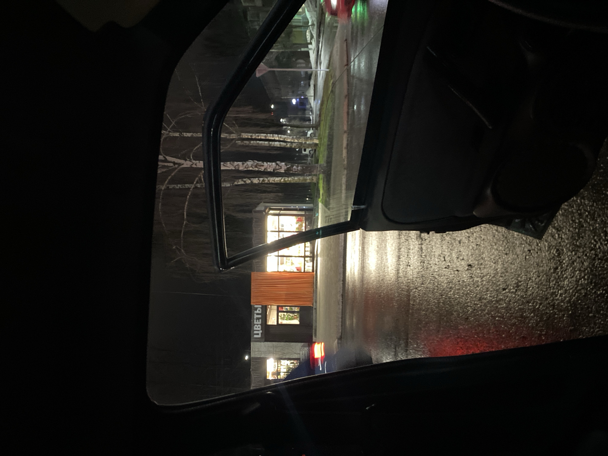 The image depicts a night scene viewed through an open car door, likely taken from the driver's seat. The location is likely Chaikovsky, Russia, as the storefront sign reads "ЦВЕТЫ," the Russian word for "flowers." The open door frames a wet street reflecting light, with a flower shop illuminated in the background. The street is lined with birch trees. The scene suggests a rainy or recently rained-upon environment. The interior of the car is dark, and details are minimal. A red tail light is visible on the street, suggesting the presence of another vehicle. The overall atmosphere is dark and urban.
