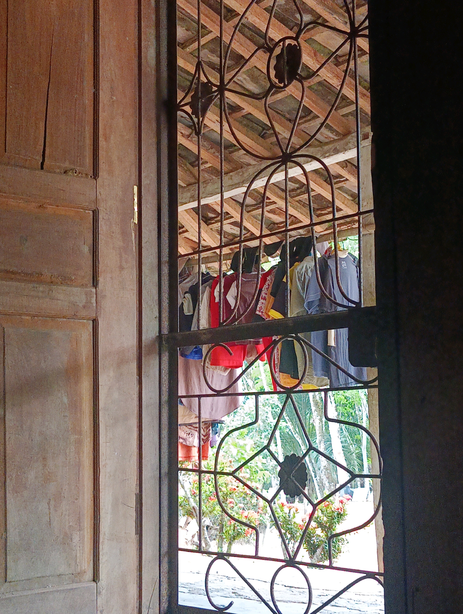 The image shows a view through a barred window in Sidorejo, Indonesia. The window has an ornate metal grate with a floral motif in the center. Outside the window, clothes are hanging on a clothesline under a wooden roof. The clothes appear to be a mix of shirts and dresses in various colors. Beyond the clothesline, lush green foliage and some red flowers are visible, suggesting a garden or natural outdoor setting. The scene is framed by a wooden door on the left and a dark, out-of-focus area on the right.