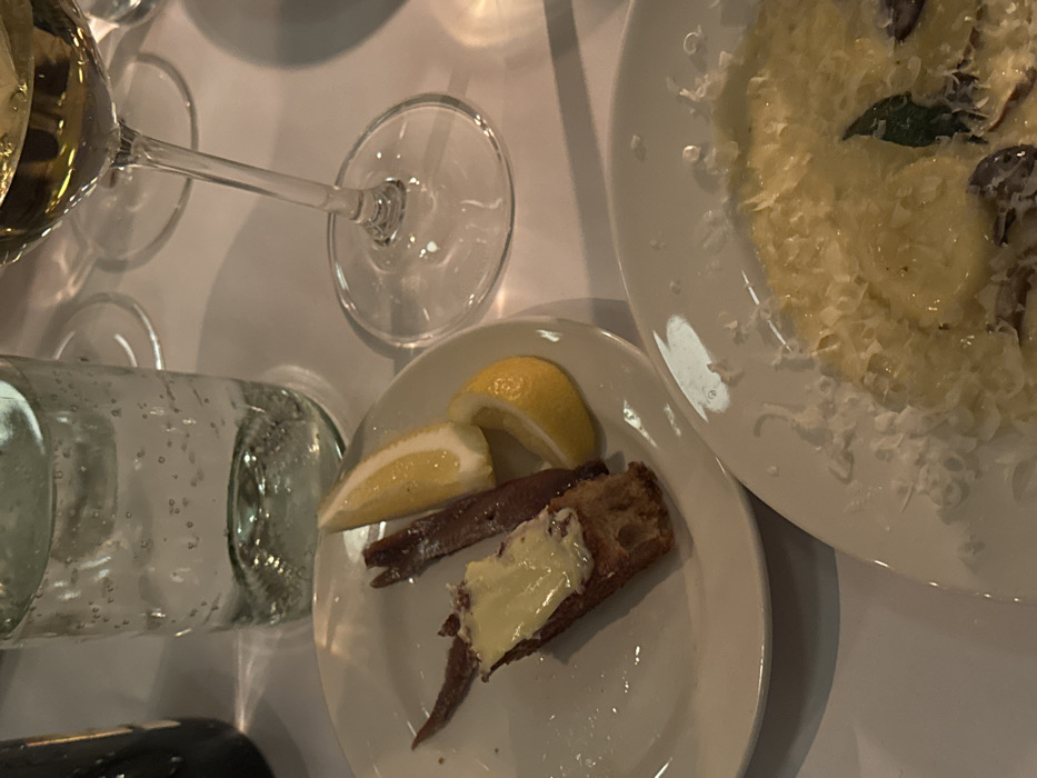 A table setting displays a plate with anchovies, lemon wedges, and bread topped with butter. Another plate holds a pasta dish sprinkled with cheese. Beside the plates are a wine glass partially filled with a light-colored liquid, an empty wine glass, and a bottle of water lying on its side. The scene is lit softly, creating a cozy ambiance for dining.