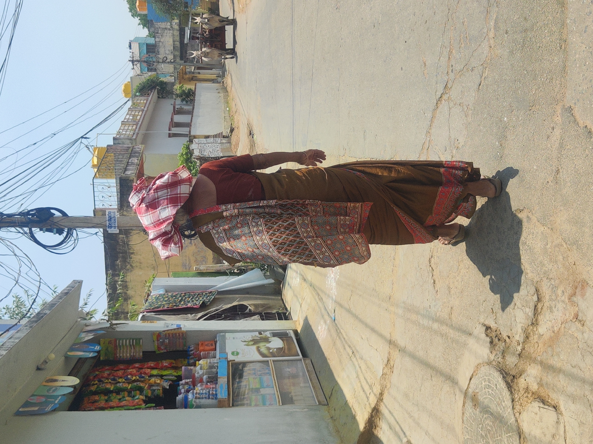 A woman in a brown saree with a red blouse and a red-and-white checkered cloth on her head walks on a concrete street in Pedda Kammavaripalli, India. She passes a small shop with snacks displayed in front. Behind her, two oxen pull a cart on the street. Above, a web of electrical wires crisscrosses the sky. The weather appears sunny and bright. The concrete street shows signs of wear and cracking.