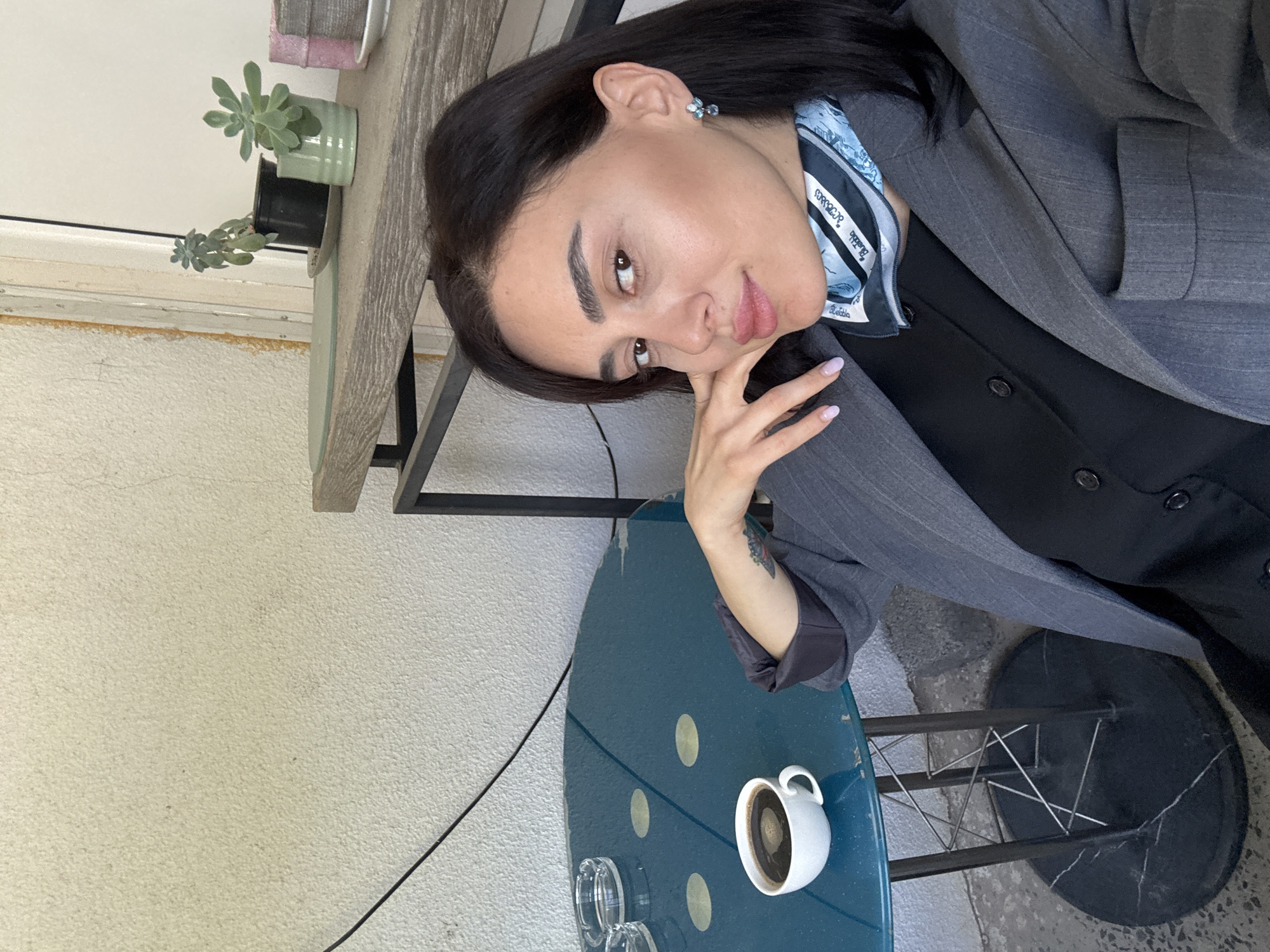 A woman is posing for a photo at a table outdoors. She wears a gray blazer over a black top, with a patterned scarf around her neck. She has dark hair, fair skin, and her hand is near her face. The table is round and teal with white polka dots. A white mug with dark liquid and two clear glass ashtrays sit on the table. There is a potted succulent plant on a shelf behind the table. The location is likely a café or outdoor seating area, potentially in Tbilisi, Georgia.