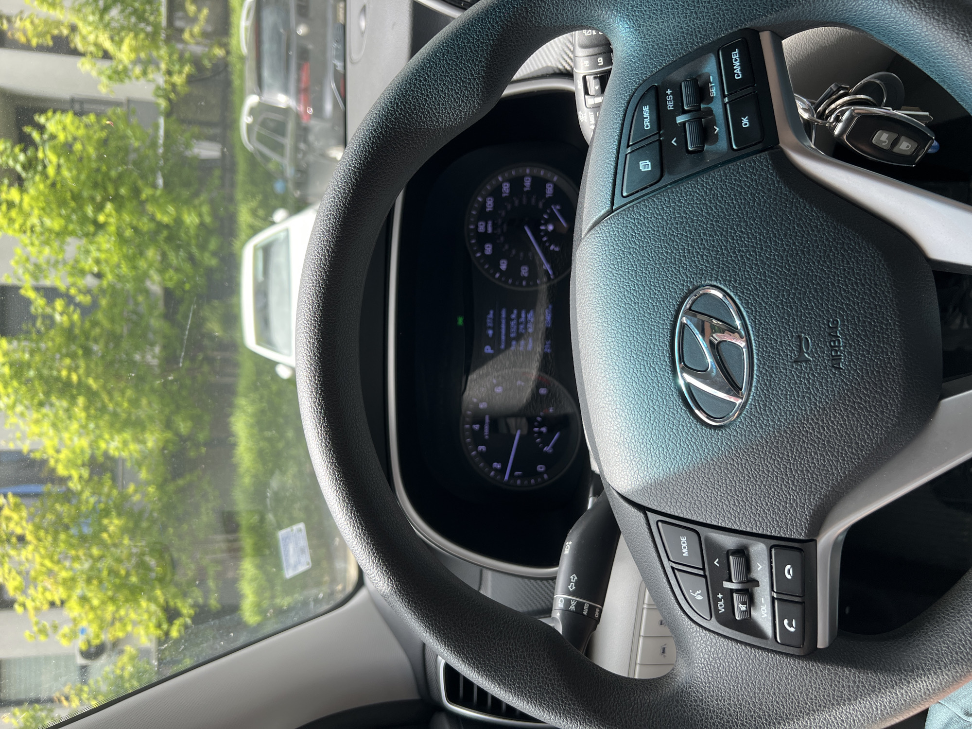 The image shows the interior of a car, focusing on the steering wheel. The steering wheel has a Hyundai logo, cruise control buttons, and volume controls. A set of keys is attached to the steering column. The car's dashboard is visible behind the steering wheel, displaying the speedometer and other gauges. Through the windshield, there is a view of green trees and buildings, indicating an outdoor setting, most probably Burgas, Bulgaria. Other vehicles are vaguely visible through the windshield as well.