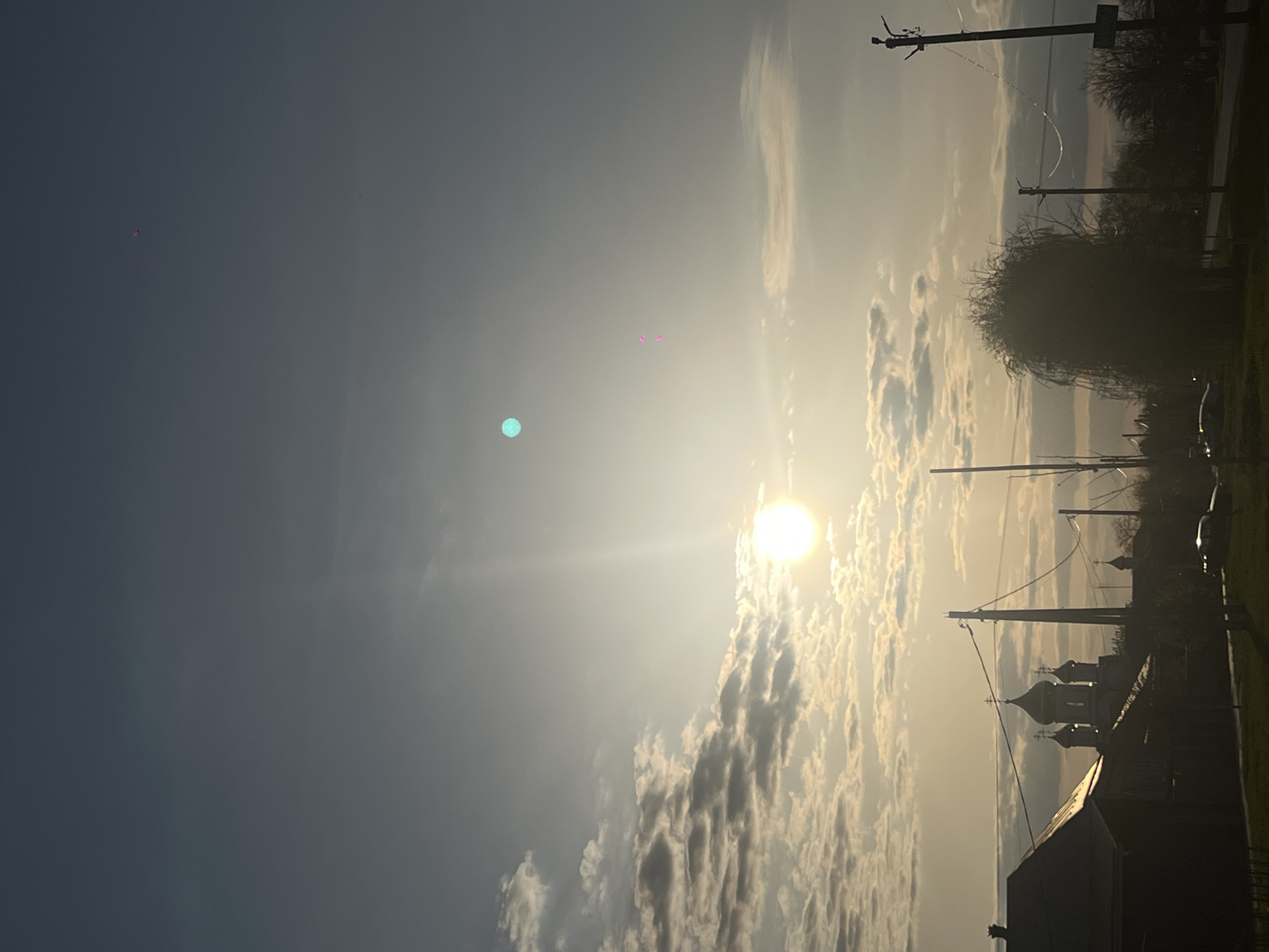 The image, when correctly oriented, presents a dramatic sunrise or sunset scene over Novopokrovskaya, Russia. The bright, luminous sun is a central feature, positioned in the middle-right of the frame, emitting strong light and creating noticeable lens flares, including distinct pink and green dots and light streaks across the sky.
The sky transitions from a darker, clear blue on the left to a brilliant, cloud-filled expanse around the sun. Clouds on the right side are illuminated with warm, golden hues, creating textured patterns against the bright backdrop.
The lower half of the image features a silhouetted landscape. Several utility poles and their wires are visible, stretching across the scene, particularly in the mid-right. Dark, bare trees and a more voluminous, bushy tree stand in silhouette against the radiant sky. A prominent architectural feature is a church with distinct onion domes, clearly visible in silhouette on the mid-right horizon, providing a strong cultural context for the location. Other indistinct building structures are also present in the dark foreground. The strong backlighting creates high contrast, emphasizing the outlines of the terrestrial subjects against the vibrant sky. There is no discernible activity or visible text.