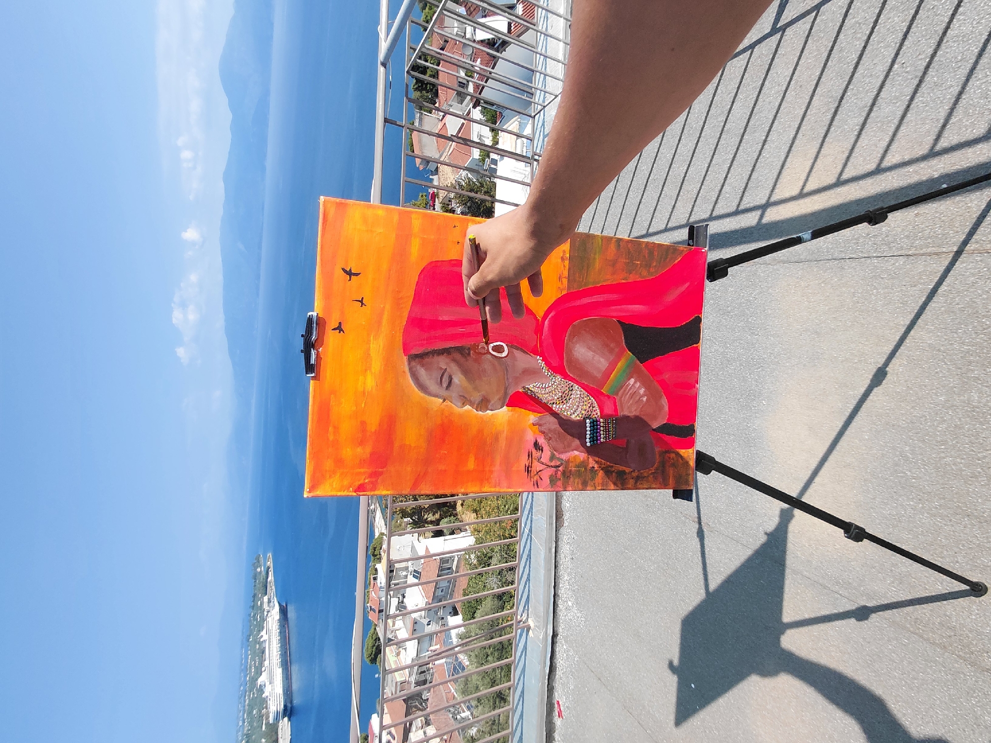 An artist's arm and hand hold a paintbrush to an oil painting on an easel. The painting depicts a woman in a red headdress and ornate jewelry against an orange background with silhouetted trees and birds. The scene is set outdoors, with a railing and a rooftop in the foreground. Beyond, a cityscape with white-roofed buildings slopes down to a blue sea. In the distance, hazy mountains form the horizon. A large cruise ship is visible docked in the bay. The location is the Municipal Unit of Aegio, Greece.