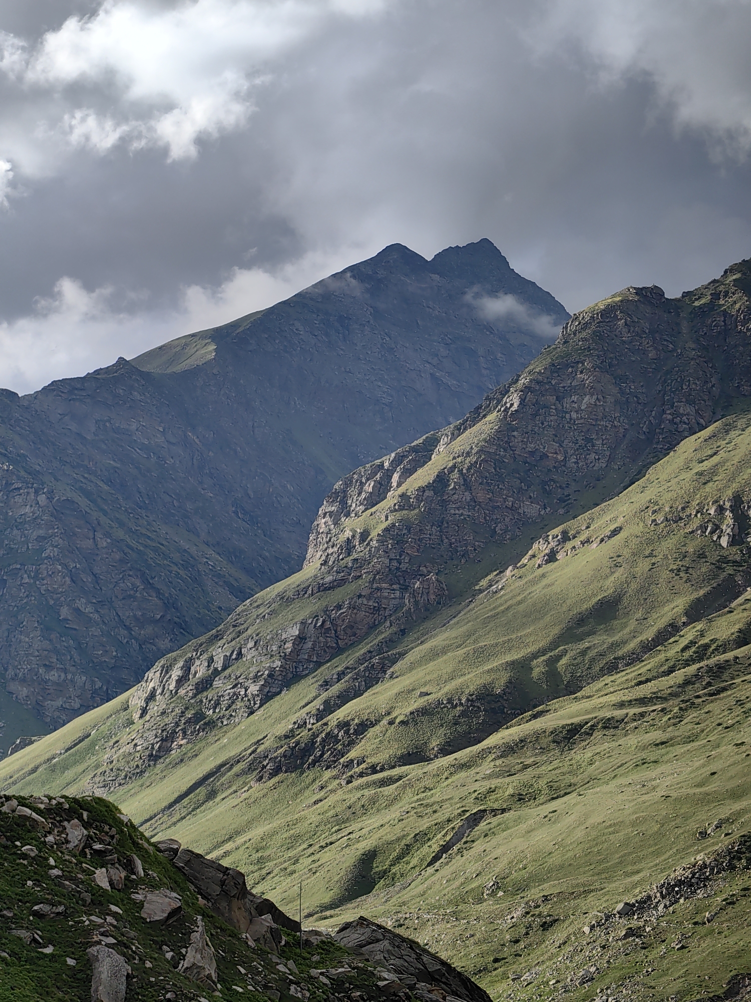 The image shows a mountainous landscape with steep, rocky slopes covered in green grass. The sky above is dramatic, with dark, heavy clouds casting shadows on the mountainside. The terrain suggests a high-altitude location, possibly in the Himalayas. The image is a photograph of the natural environment. The location is specified as Burwa, India.