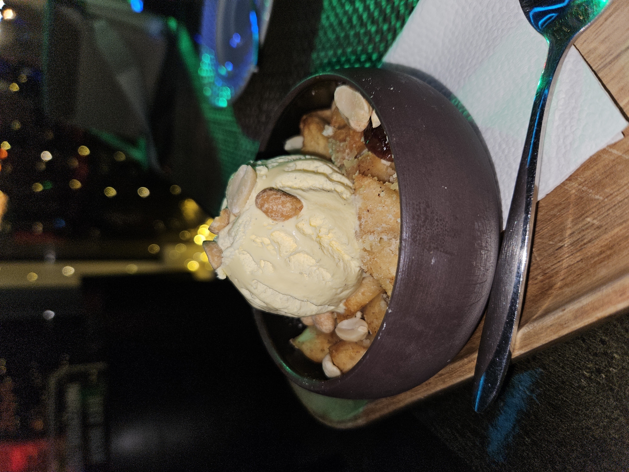 A close-up, slightly tilted, indoor shot shows a dessert in a brown bowl, placed on a small wooden tray with a folded white napkin and a spoon, likely in Unknown, Unknown. The dessert features a scoop of pale yellow ice cream atop what seems to be a bread pudding or crumble, garnished with sliced almonds and whole nuts. The bowl is dark brown and has a smooth, matte finish. The wooden tray has a natural wood grain. The spoon appears to be stainless steel, catching reflections of blue and green lighting. The napkin is neatly folded beneath the bowl. The background shows blurry lights in red, yellow, blue, and green, suggesting a restaurant or bar setting. The overall lighting is low, emphasizing the dessert.