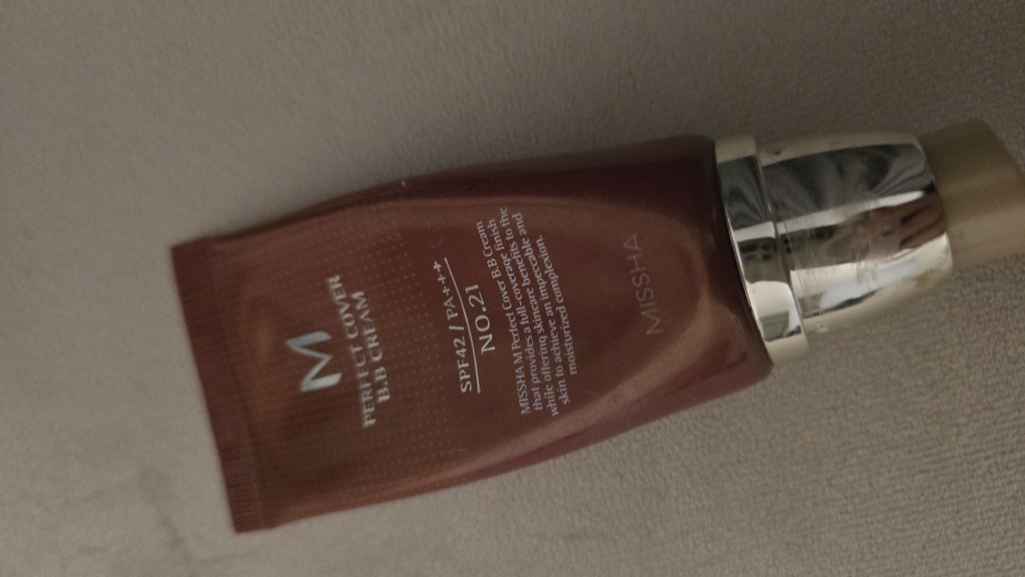The image shows a tube of Missha M Perfect Cover BB Cream, shade No. 21, resting on a textured, light-colored surface. The tube is a reddish-brown color with white text. The top portion of the tube features a large white "M" logo, followed by "PERFECT COVER B.B CREAM". Below that, it reads "SPF42/PA+++ NO.21". Further down, there is a detailed description of the product: "MISSHA M Perfect Cover B.B Cream that provides a full-coverage finish while offering skincare benefits to the skin to achieve an impeccable and moisturized complexion." At the bottom of the tube, the brand name "MISSHA" is printed. The bottom of the tube has a silver cap. The overall scene is a close-up of the product itself, with no indication of its location within the city of Batumi, Georgia, or any people, actions, or interactions.