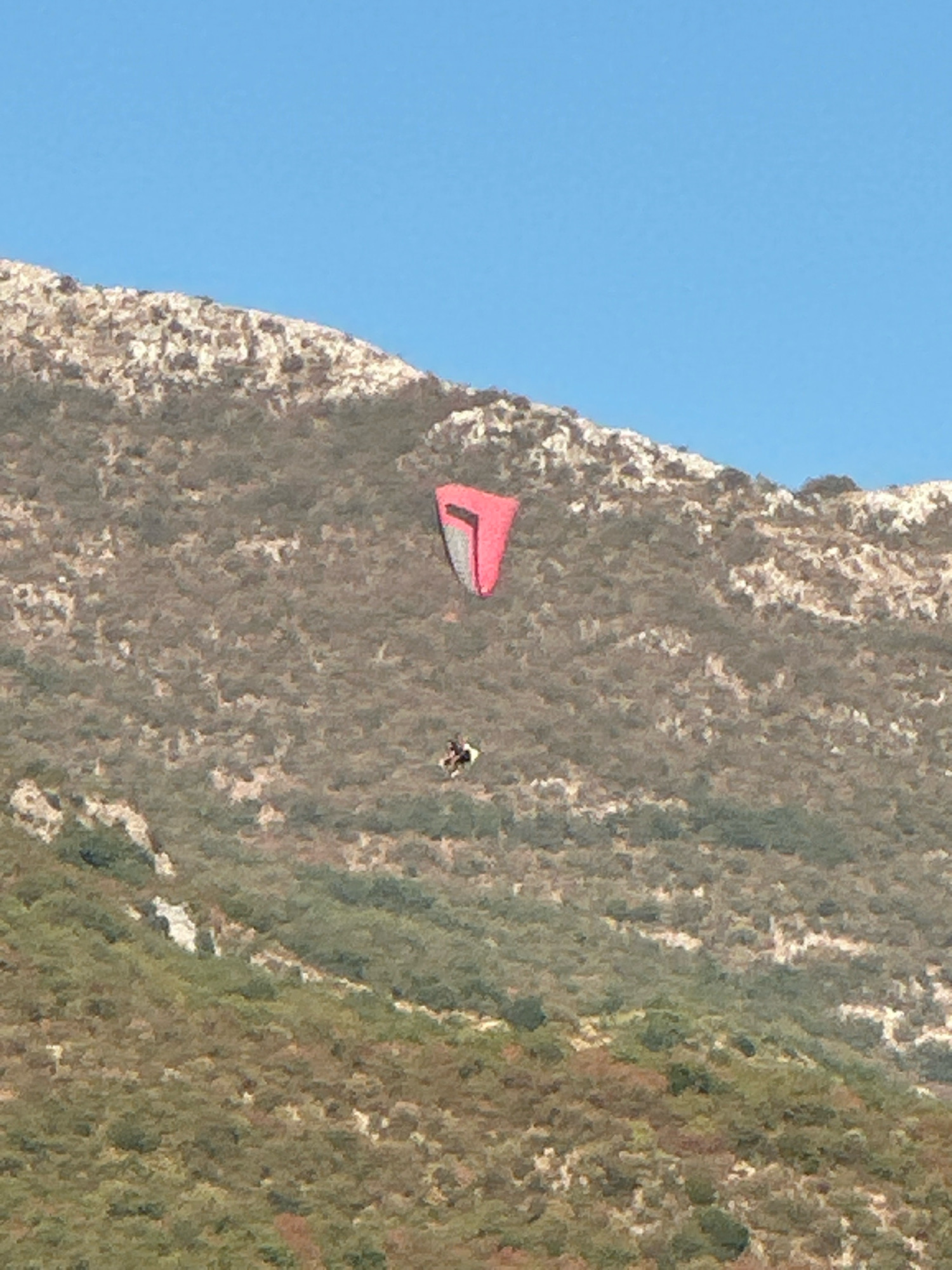 An outdoor, high-angle, medium shot shows a paraglider in mid-flight against a clear blue sky and a barren, rocky hillside. The paraglider, colored pink and gray, is positioned in the upper-middle section of the frame, appearing to be descending. Below the paraglider, a small figure, presumably the paraglider pilot, is visible, wearing dark clothing and suspended in a harness. The hillside is covered with sparse vegetation and rocks, with a patch of greener trees visible in the lower portion of the image. The scene is illuminated by bright sunlight, casting shadows on the terrain. The location is identified as Boreti, Montenegro.
