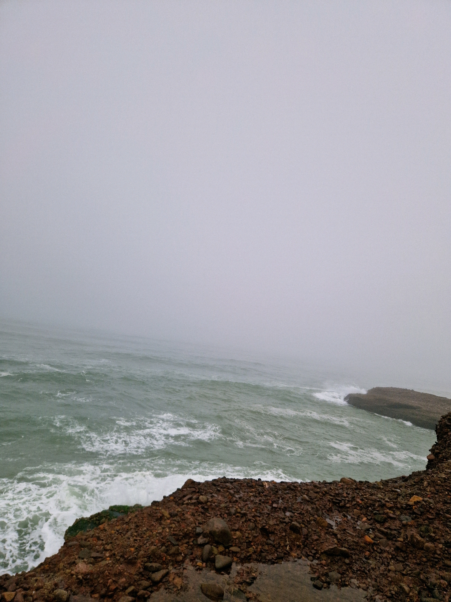 The image depicts a rugged, rocky coastline with the ocean meeting the shore. The sea is a turbulent, greenish-blue with whitecaps, suggesting a moderate to rough surf. The sky is overcast and foggy, creating a muted, diffused light and obscuring the horizon. The foreground is dominated by a steep bank of dark brown and reddish-brown rocks, some of which are quite large. There is some sparse green vegetation clinging to the rocks in places. In the distance, a rocky outcrop juts out into the sea. The scene is devoid of any human presence or man-made structures. The location is Foum Assaka, Morocco.
