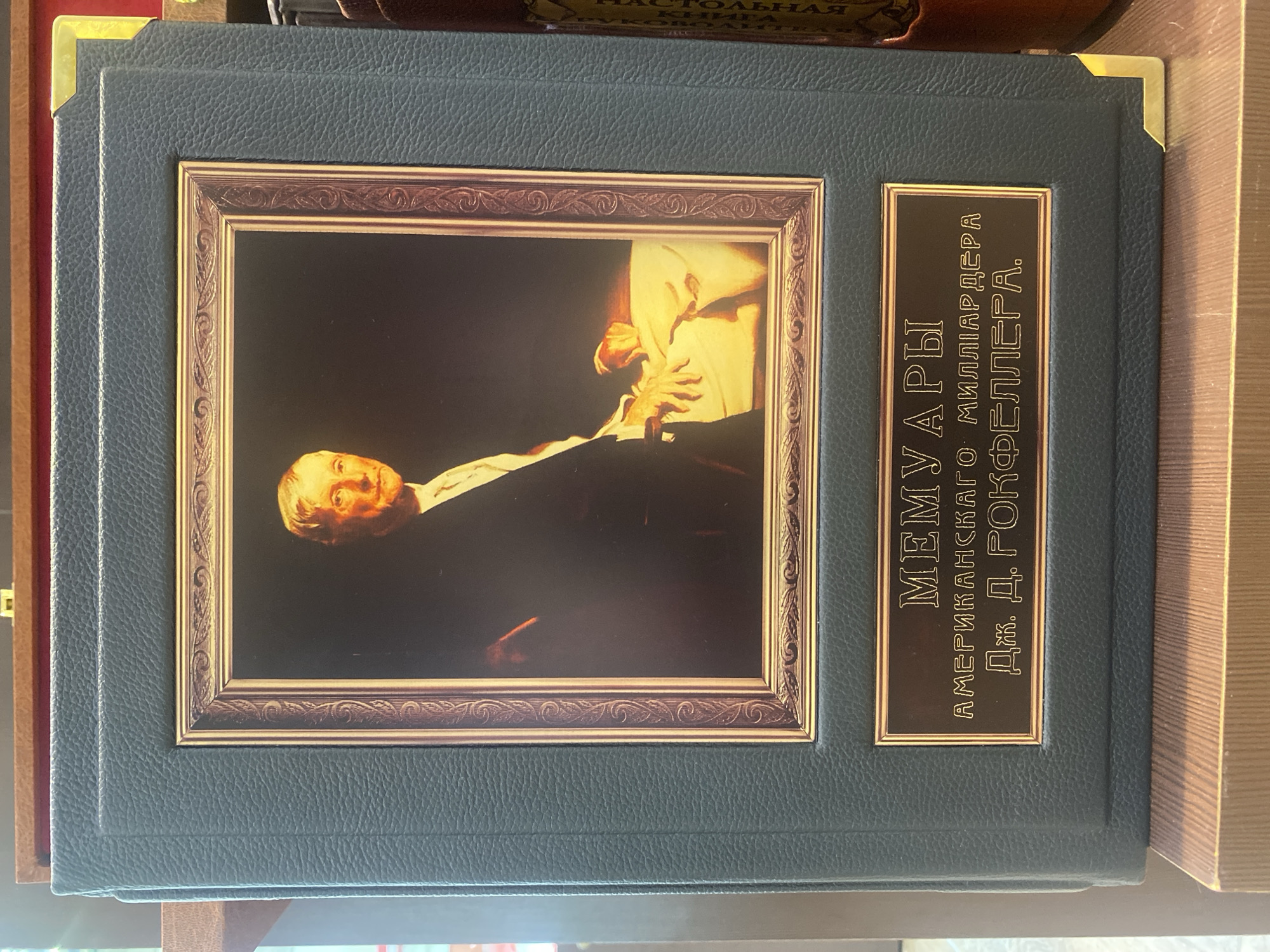 The image shows a dark teal bound book with a portrait of an elderly white man in a suit inside a decorative gold frame. Below the portrait is a brass plaque with text in Russian. The book is resting on a wooden surface, likely a shelf, in what appears to be an indoor setting. The overall presentation suggests it is a collection or a special edition of a book.