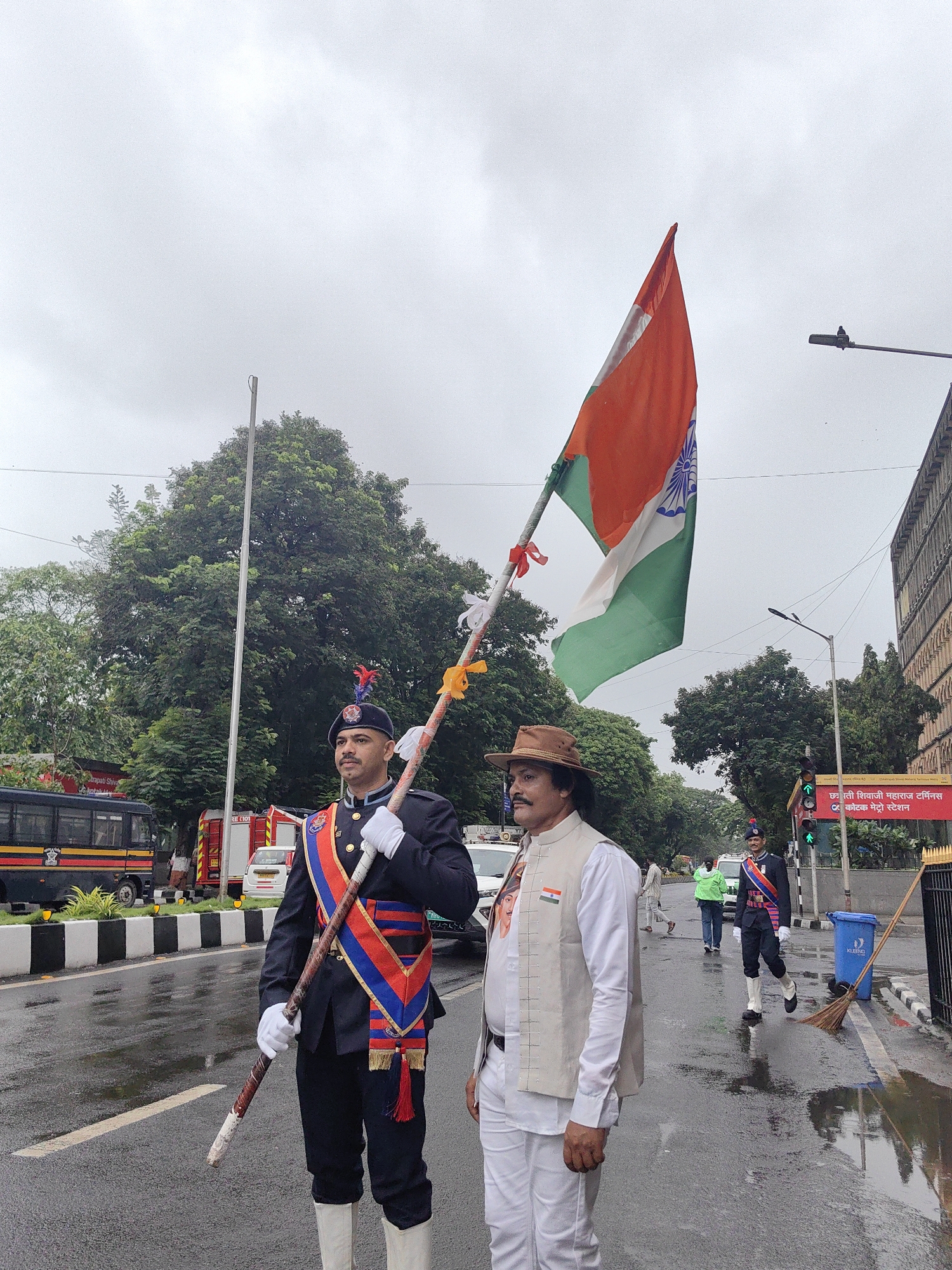 Two men are in the foreground of the image, with an Indian flag held by the man on the left. The man on the left is wearing a dark blue uniform with red and blue trim, a dark blue beret with a badge, white gloves, and white boots. He is holding a long pole with red and yellow ribbons. The man on the right is wearing a white shirt, a tan vest adorned with a small Indian flag, and white pants. He has a hat on his head and a mustache. They are standing on a wet road, suggesting it has recently rained.
In the background, there are trees, a large building, a bus, and what appears to be fire trucks. There are also streetlights and traffic signals visible. One traffic signal shows a green light. A blue trash can and a broom are to the right of the men. In the background, a banner with Marathi text is visible on a building. The text on the banner reads "छत्रपती शिवाजी महाराज टर्मिनस" which translates to "Chhatrapati Shivaji Maharaj Terminus" and "कळोबा मेट्रो स्टेशन" which translates to "Kaloba Metro Station." This indicates the location is near Chhatrapati Shivaji Maharaj Terminus in Mumbai, India. The overall scene depicts an outdoor event, possibly a parade or ceremony, on a cloudy day.