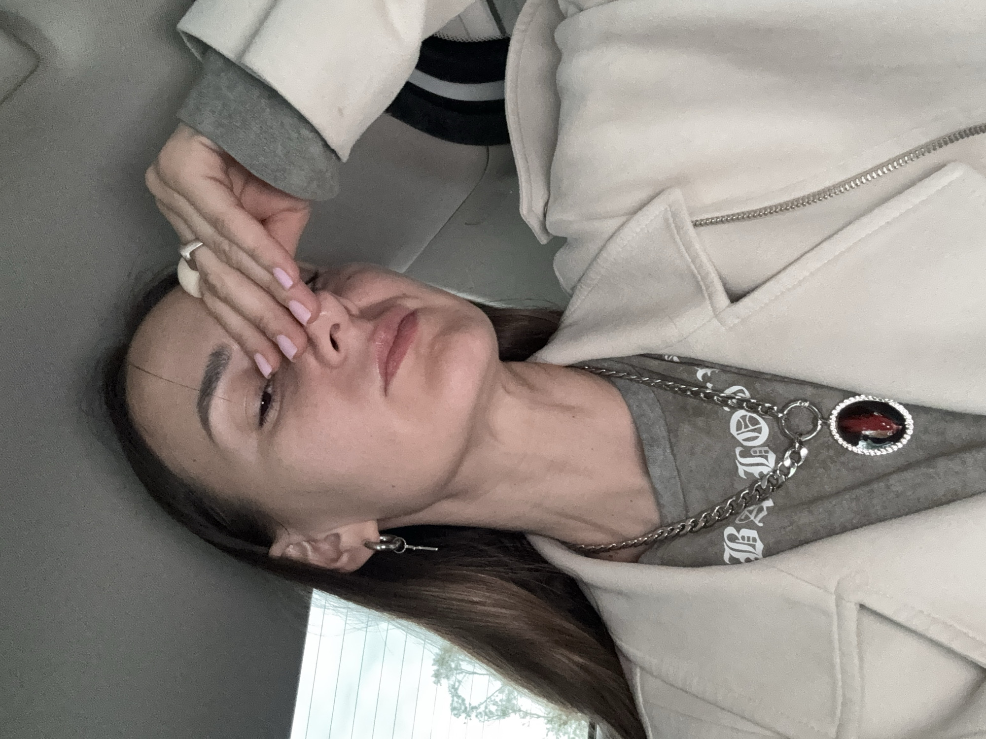 A woman is seated indoors, possibly in a car given the blurred window and interior panel in the background. She is wearing a light-colored coat or jacket over a gray t-shirt with some white letters. She has a silver necklace with a pendant. Her hand is covering part of her face near her nose. She is wearing a silver ring on her finger and has light pink nail polish. The location of Sochi, Russia is not discernible from the image.