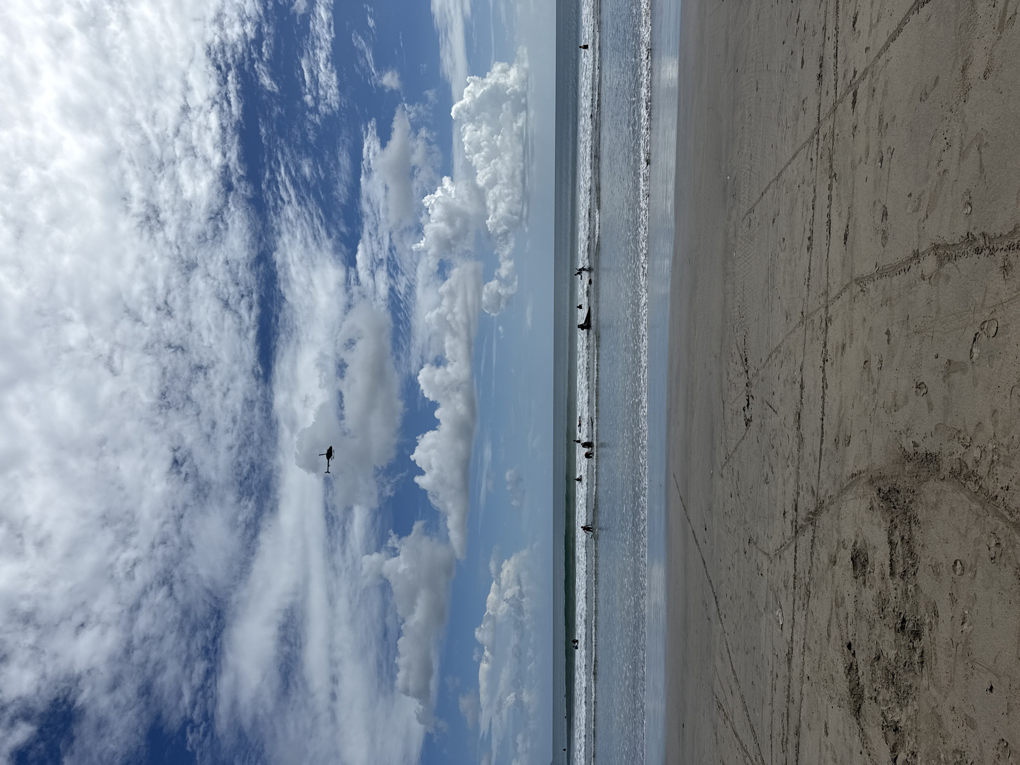 The image displays a wide, vibrant daytime scene of a beach in Kuta, Indonesia, with the sky, ocean, and sand vertically layered.
The upper half of the image is dominated by a vast sky. It features a bright blue expanse broken by numerous white clouds, ranging from large, fluffy cumulus clouds to more delicate, streaky cirrus formations. Against this bright background, the dark silhouette of a helicopter is prominently visible towards the upper left portion of the sky.
The middle section of the image shows the ocean meeting the shore. Gentle waves are breaking close to the beach. Several dark, silhouetted figures of people are scattered within the shallow water, appearing to be engaged in water activities like swimming, wading, or possibly learning to surf.
The lower part of the frame consists of the sandy beach. The sand is damp near the water's edge and shows various marks, including distinct footprints and tire tracks, suggesting recent human and possibly vehicle activity. The sand's texture appears varied, with some darker, more disturbed areas.
The scene depicts a lively coastal environment under a bright, clear sky with moderate cloud cover, indicating pleasant weather conditions. The presence of people in the water and tracks on the sand implies a recreational setting. There is no visible text in the image.