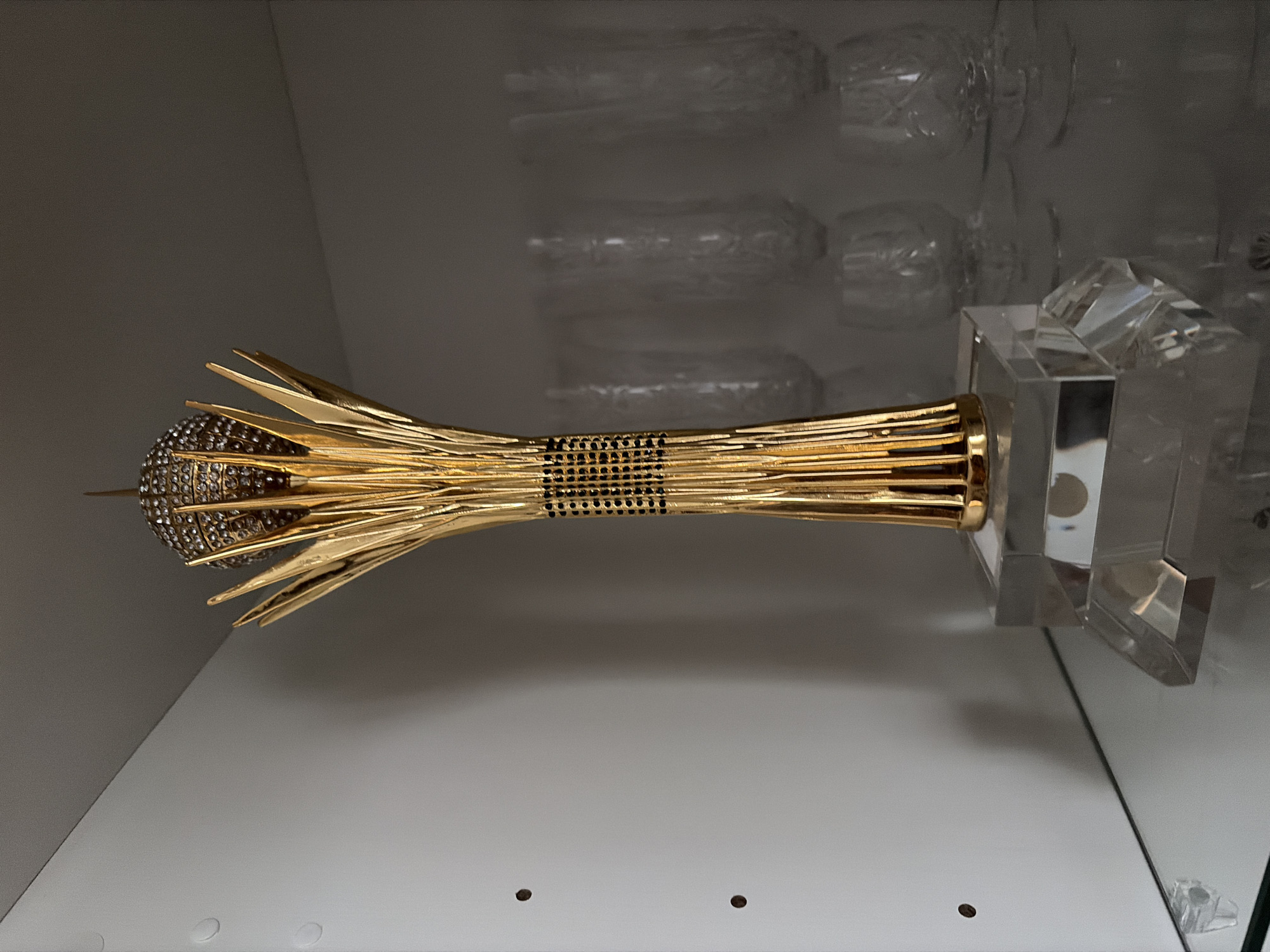 A golden trophy, adorned with clear rhinestones at its apex, stands on a clear acrylic base. The trophy's stem features a band of black and gold square embellishments. It is positioned on a glass shelf within a white cabinet. Behind the trophy, several clear, etched glasses are visible on another glass shelf. The background consists of a white cabinet wall with two small, dark circular indentations. The overall setting appears to be an indoor display or storage space. This trophy resembles the "Golden Person" award from Kazakhstan, a prestigious prize awarded annually in Astana (now Nur-Sultan) for achievements in various fields, such as sports, culture, and business.